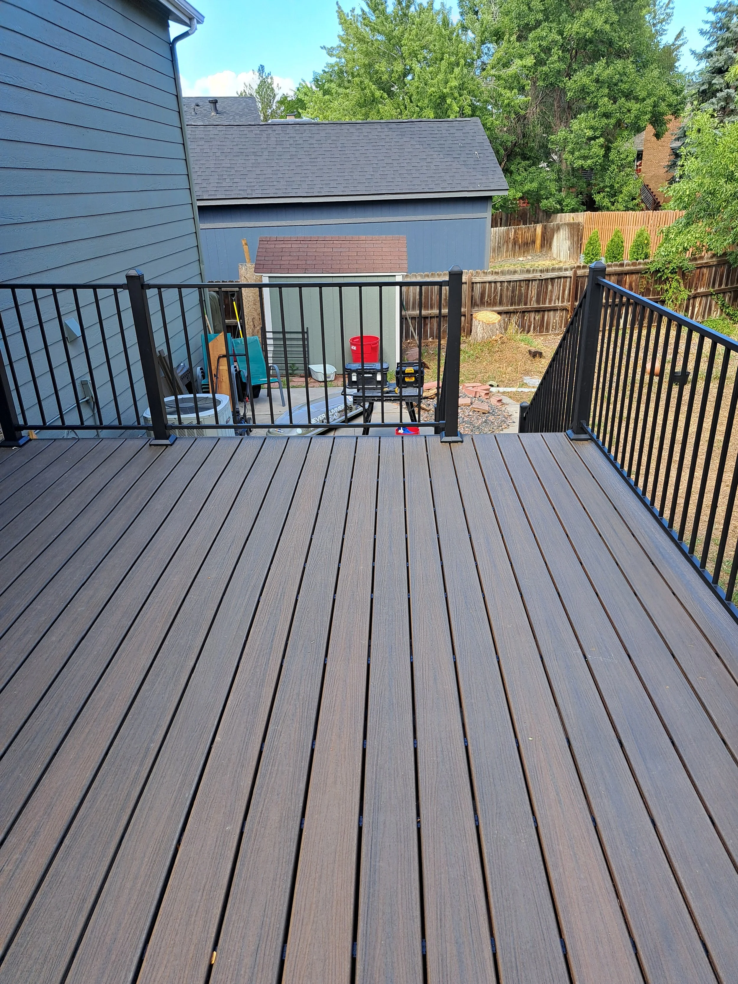 View of a backyard deck with black railing, overlooking a yard with various tools, a small shed, and trees behind a wooden fence.