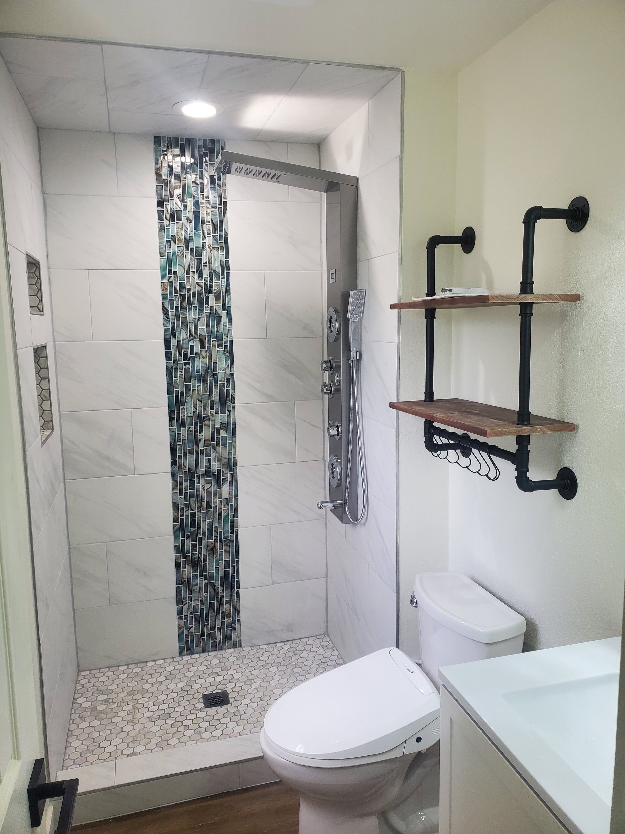 A modern bathroom with a walk-in shower featuring large white tiles, a vertical strip of multicolored mosaic tiles, and a ceiling with a recessed light. There is a white toilet, a small white vanity with a sink, and a black pipe-style wall-mounted shelf above the toilet with two wooden shelves and black pipe brackets.