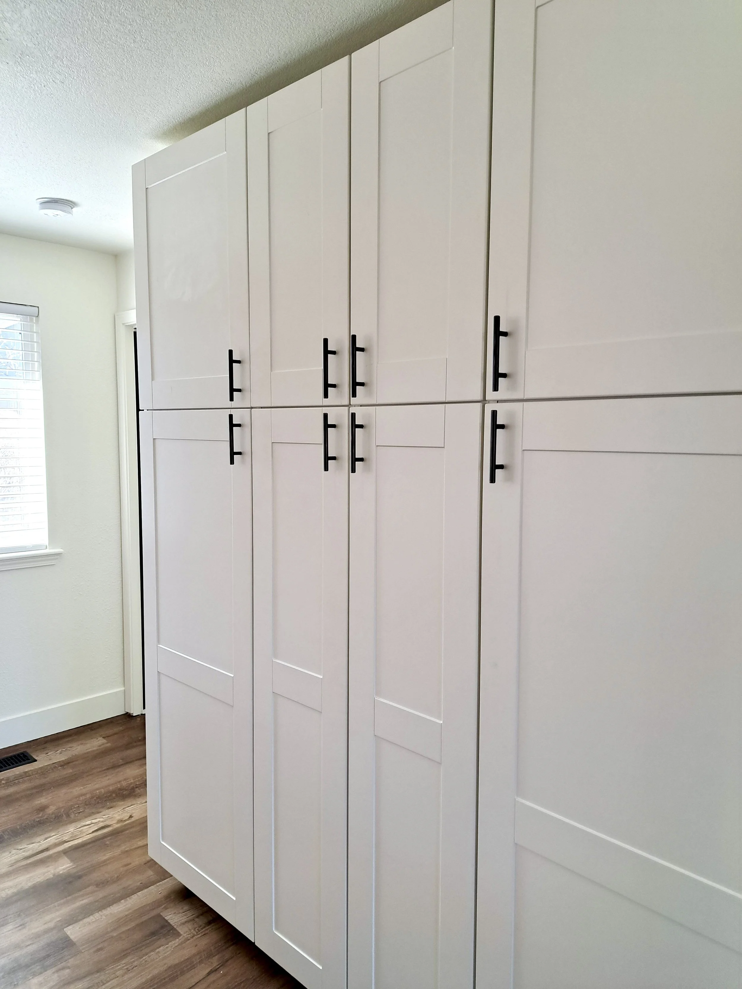 White built-in kitchen cabinets with black handles, wood flooring, and a window with blinds.