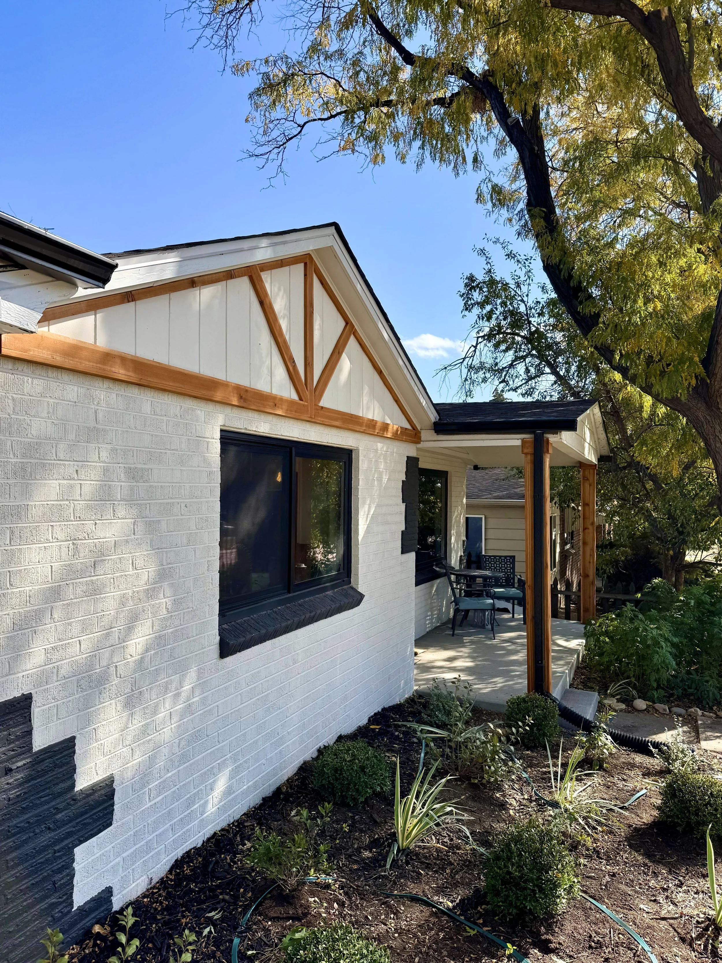 View of a white brick house exterior with a black window and an unfinished gable trim with exposed brown wooden framing, a small porch with black chairs, and a garden with small green bushes, a large tree, and clear blue sky.