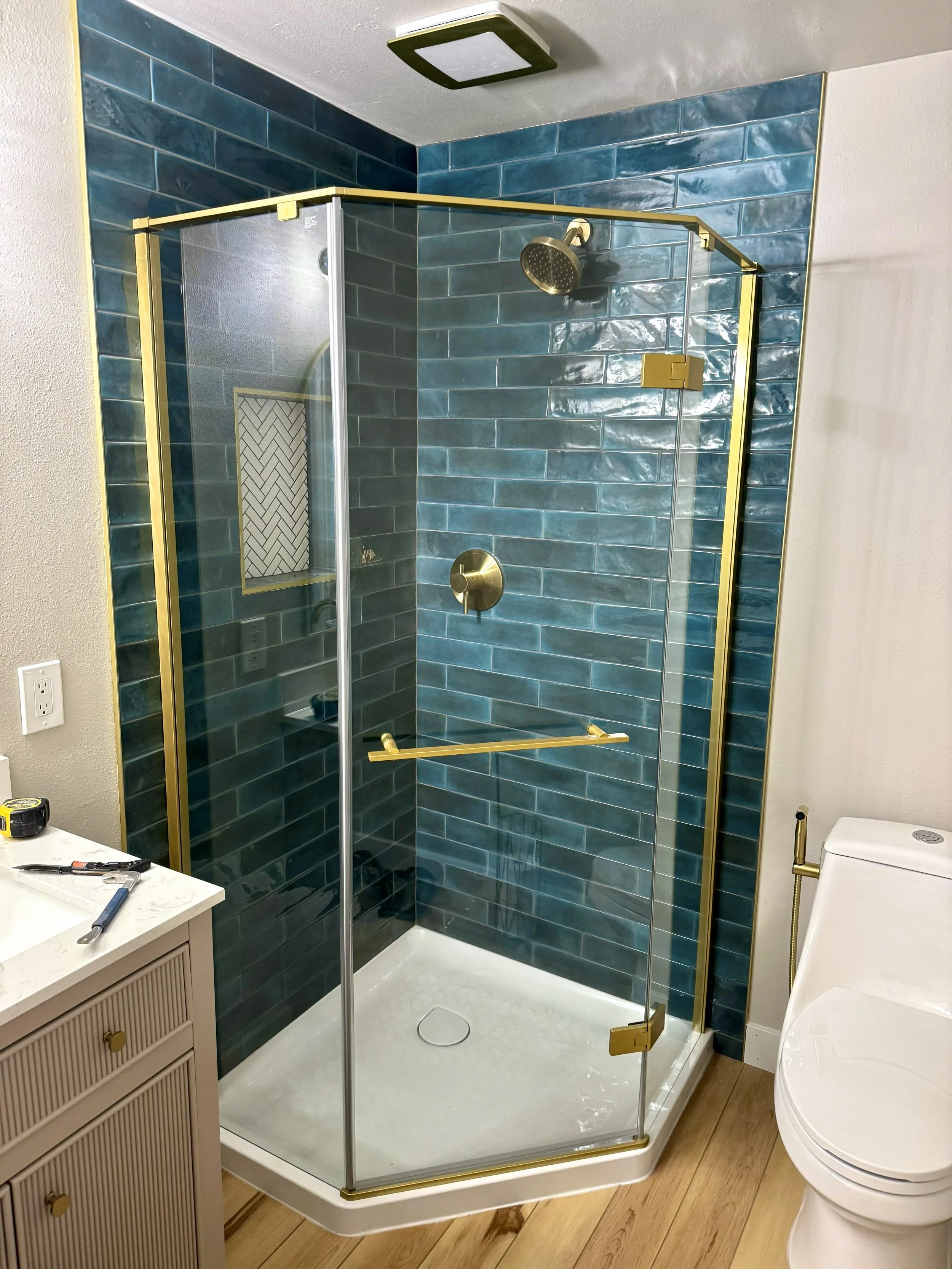 A modern bathroom shower with teal subway tiles, glass door with gold hardware, and relaxing fixtures, next to a white toilet and beige vanity.