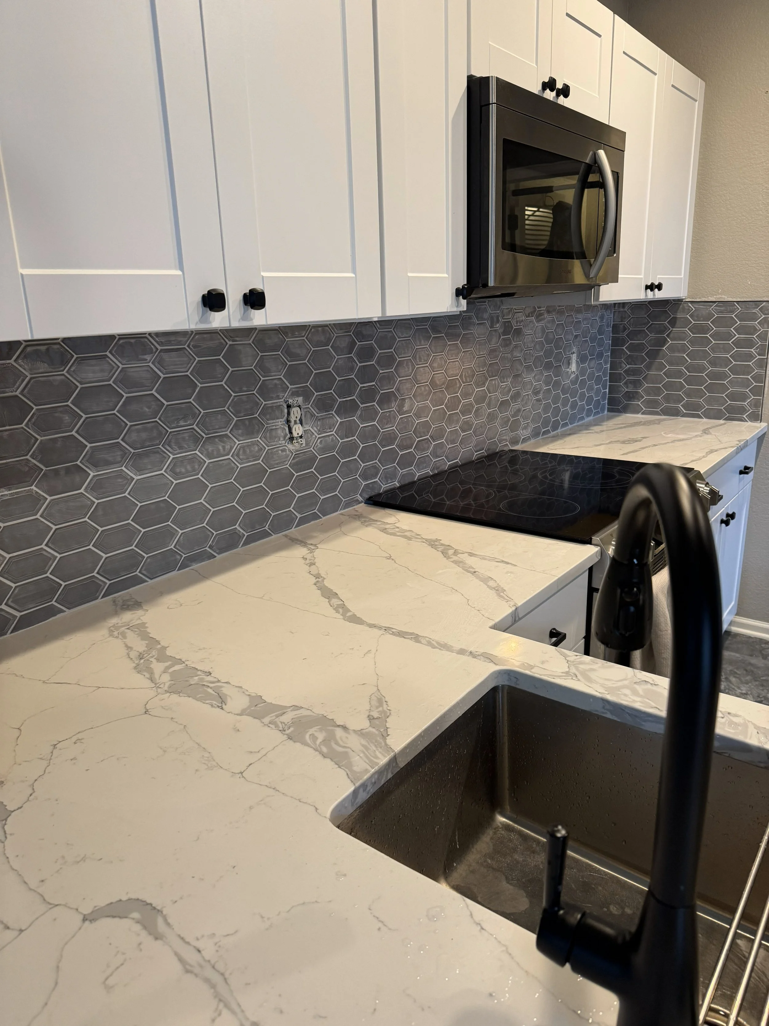 Modern kitchen with white cabinets, gray hexagonal tile backsplash, marble-patterned countertop, stainless steel microwave, black stovetop, and black faucet over a stainless steel sink.