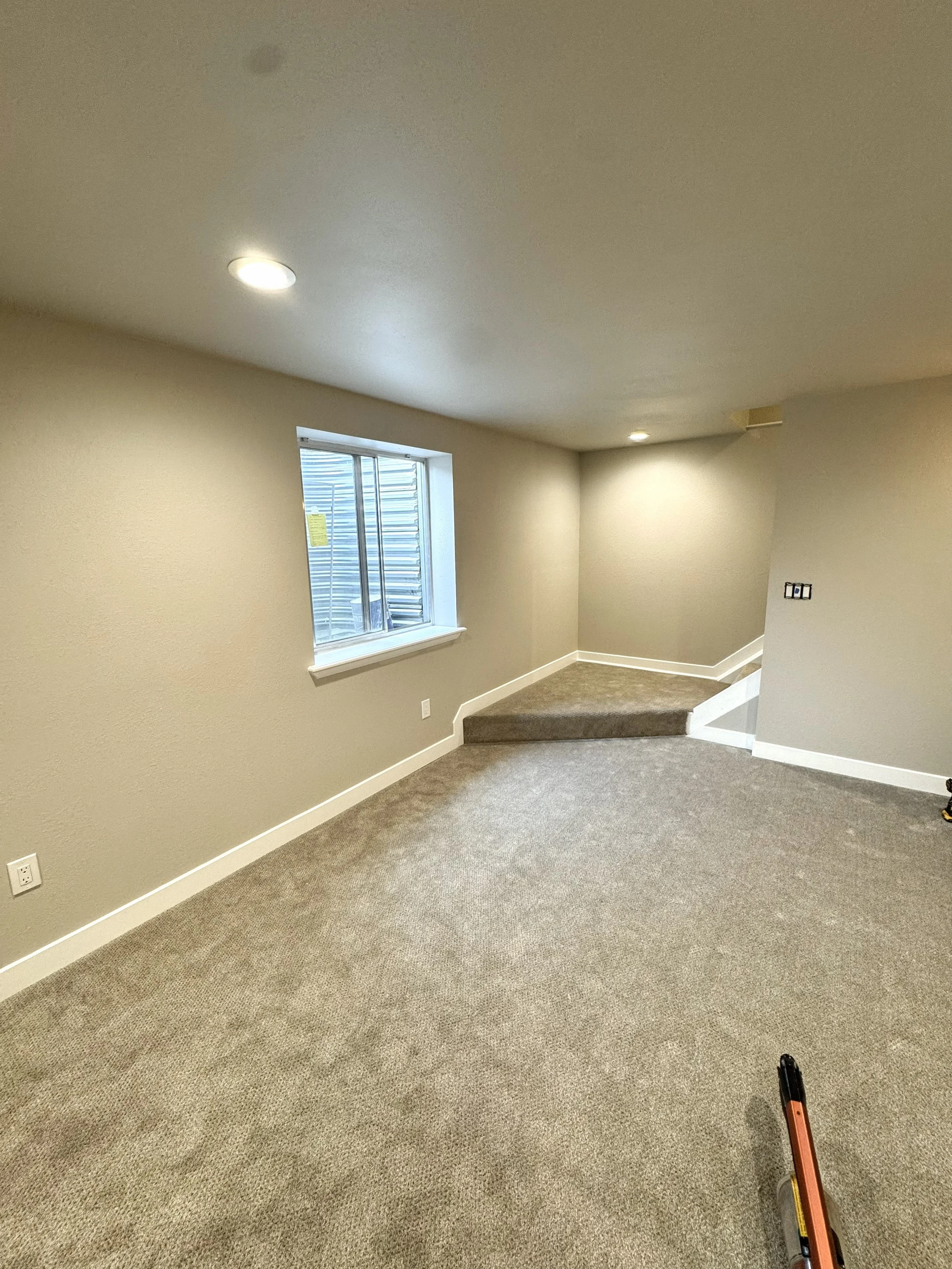 Empty room with beige carpet, beige walls, a small window, and recessed lighting. A staircase with carpeted steps leads to an upper level, and there's a yellow level tool on the floor.