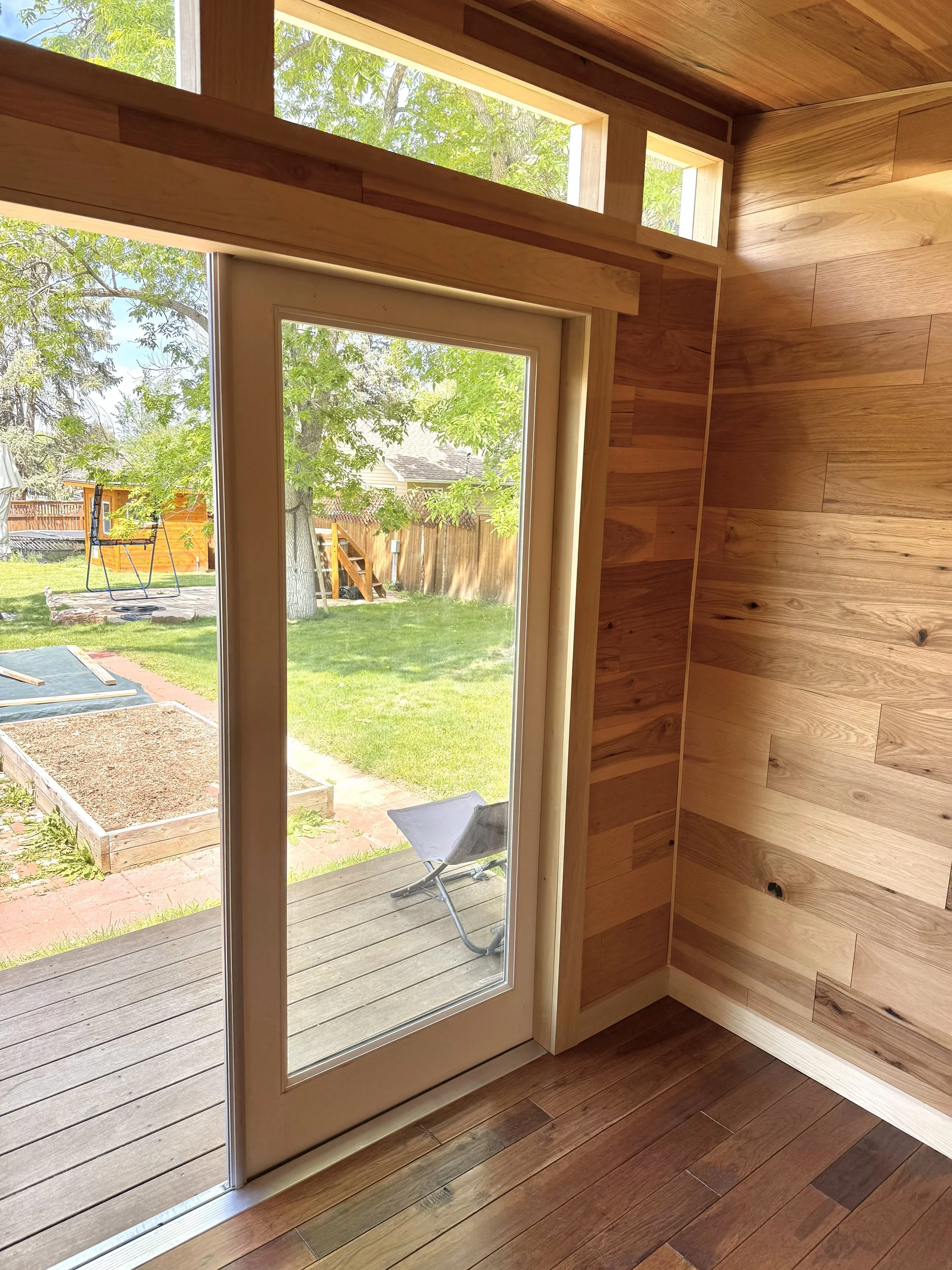 A room with wooden walls and floor, a glass door leading outside to a backyard with grass, trees, a swing set, and a lounge chair visible through the window.