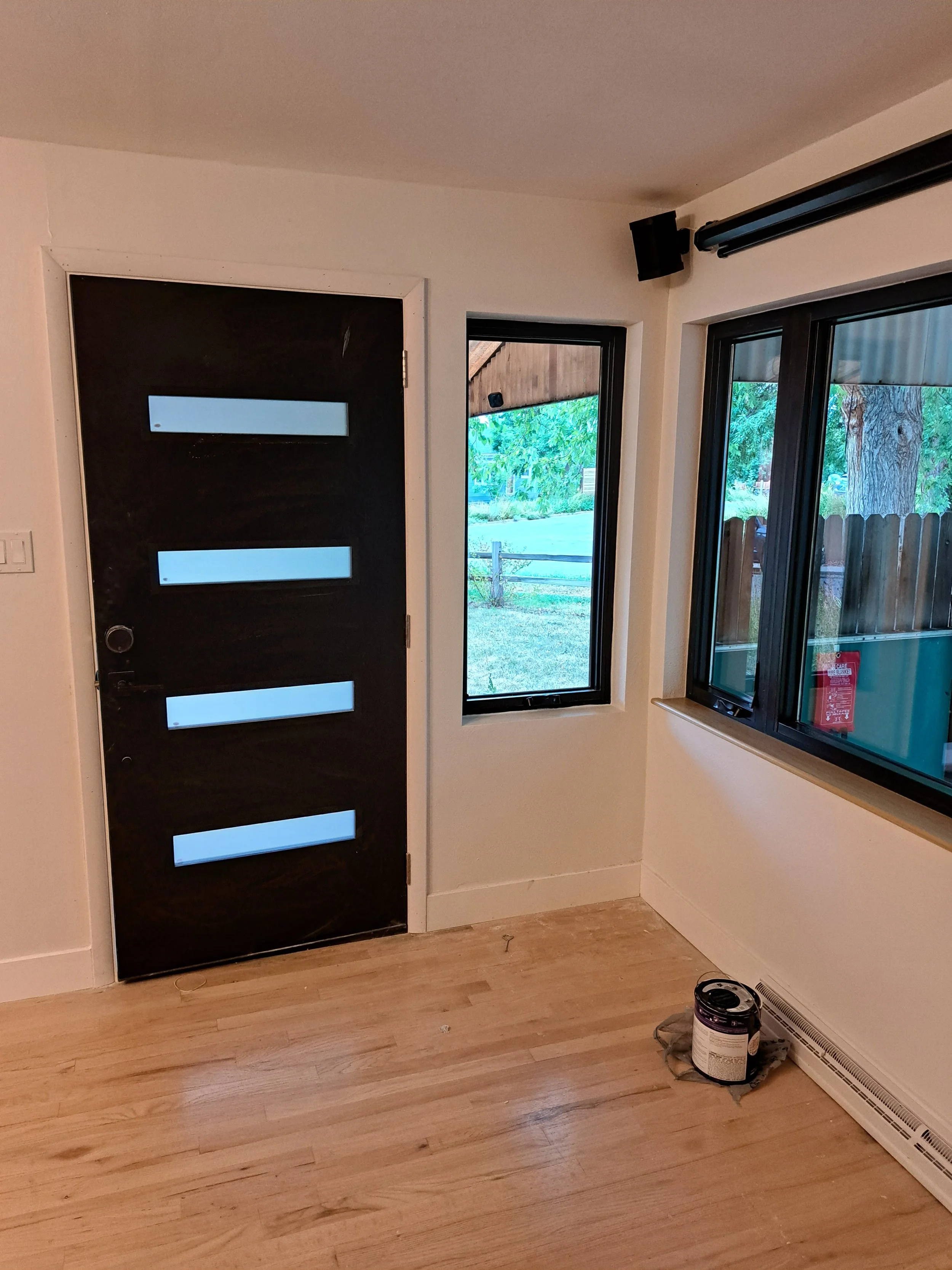 View of a small room with a black door featuring four horizontal windows, two windows on the adjacent wall showing a yard with trees, and a can of paint on the wooden floor near a wall heater.