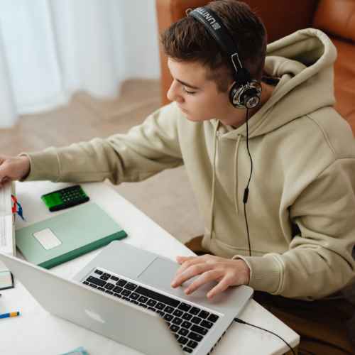 Young man wearing headphones using a laptop at a white desk with a green notebook, calculator, and other stationery.