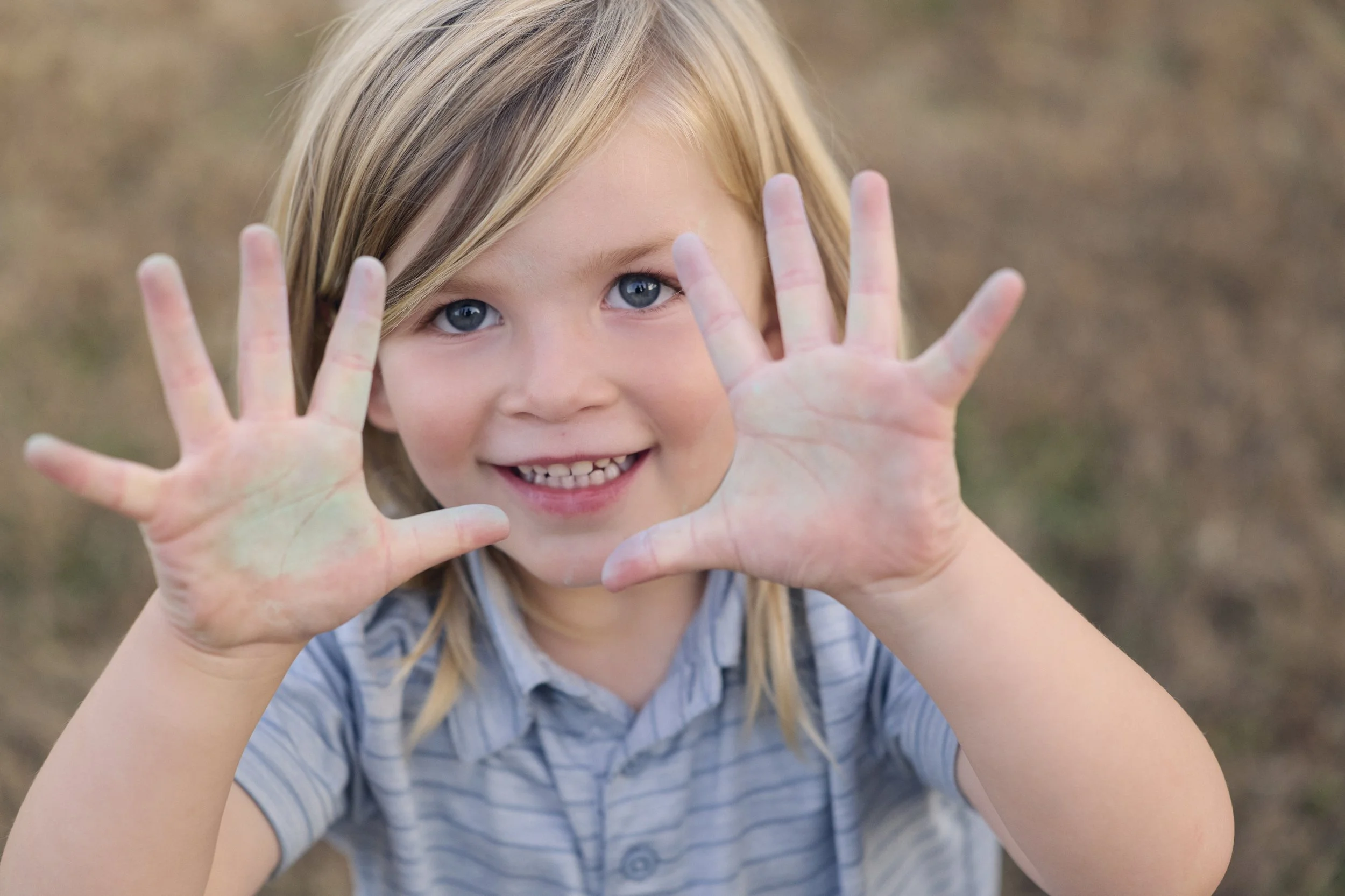 Young blonde-haired girl with blue eyes smiling and holding her hands up toward the camera, outdoors in a natural setting.
