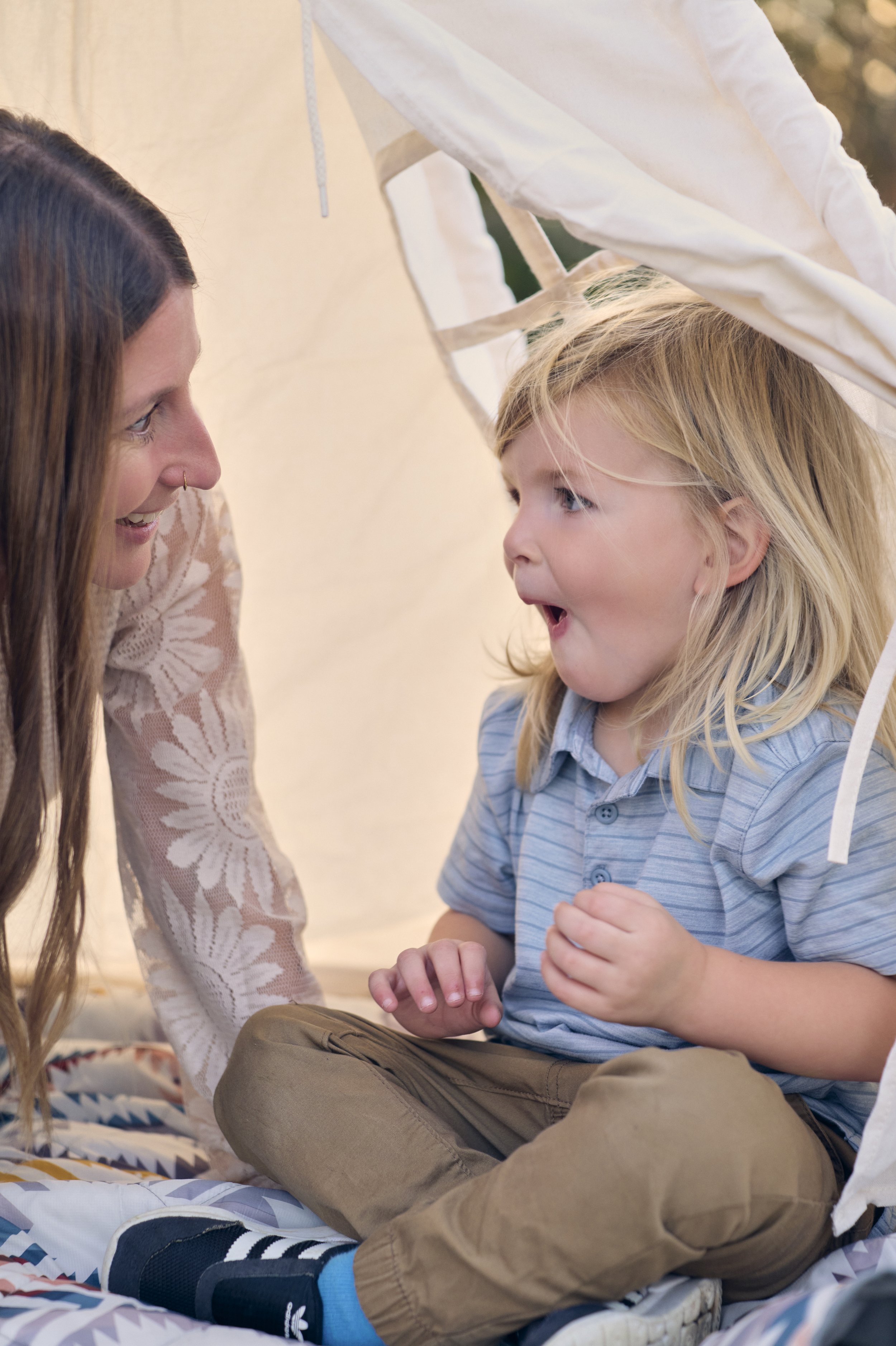 A woman and a young boy are inside a tent, looking at each other and smiling, sharing a joyful moment.