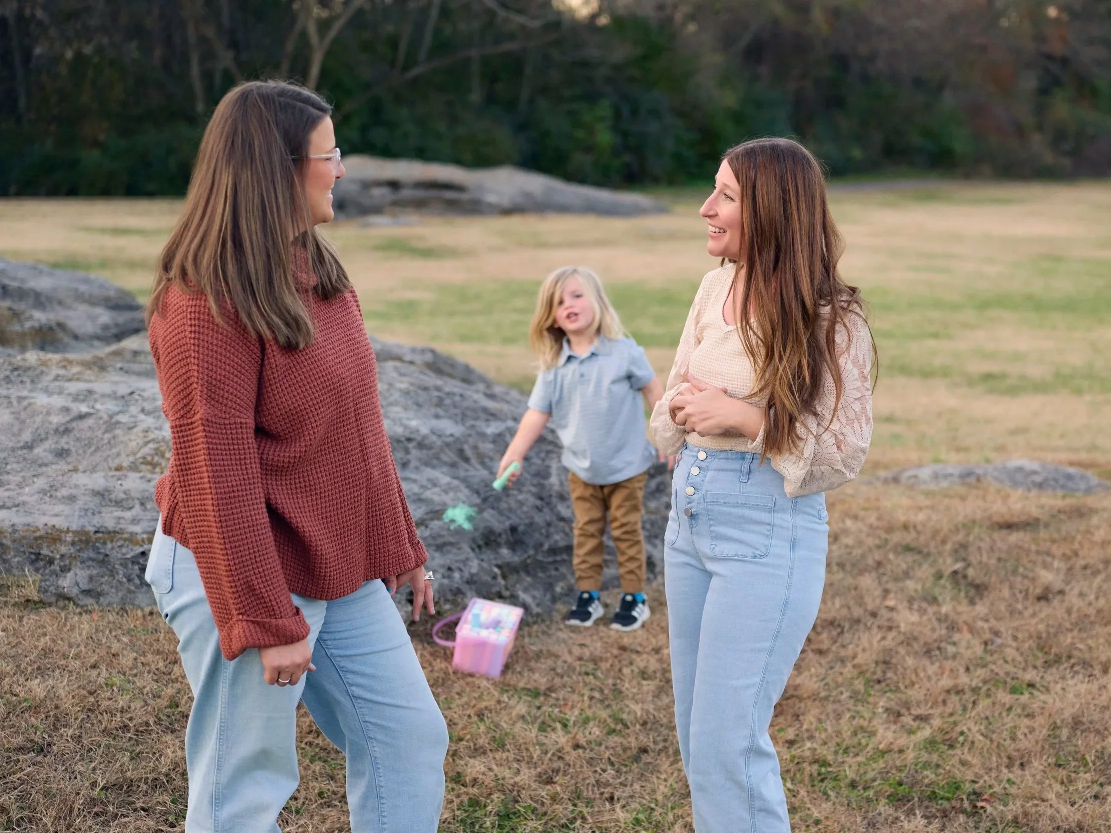 Two women standing outdoors in conversation with a young girl in the background playing with chalk near rocks and grass.