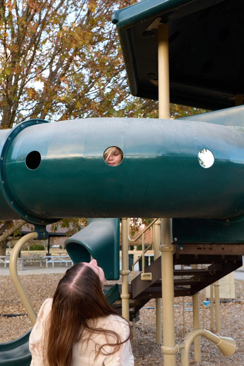 A young girl with long brown hair looking up at a boy in a playground tube. The boy's face, partially visible, is inside a green tube, with trees and a park in the background.