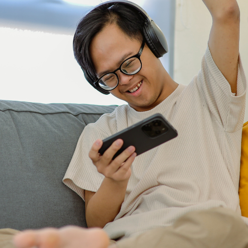 A person sitting on a gray couch, smiling and looking at a smartphone, wearing headphones and glasses.