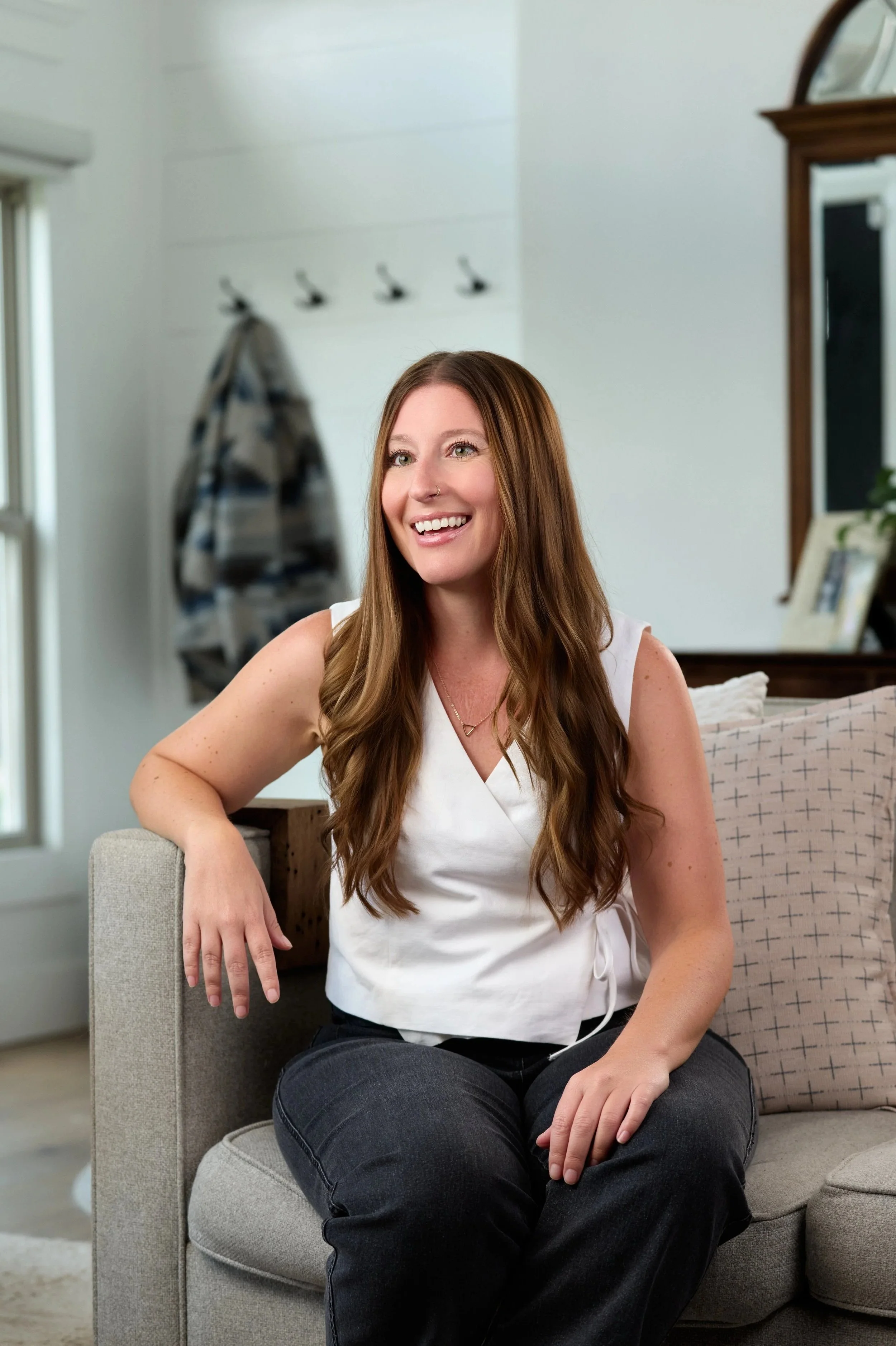 A woman with long brown hair, wearing a white sleeveless top and dark jeans, sitting on a gray couch, smiling inside a bright living room.