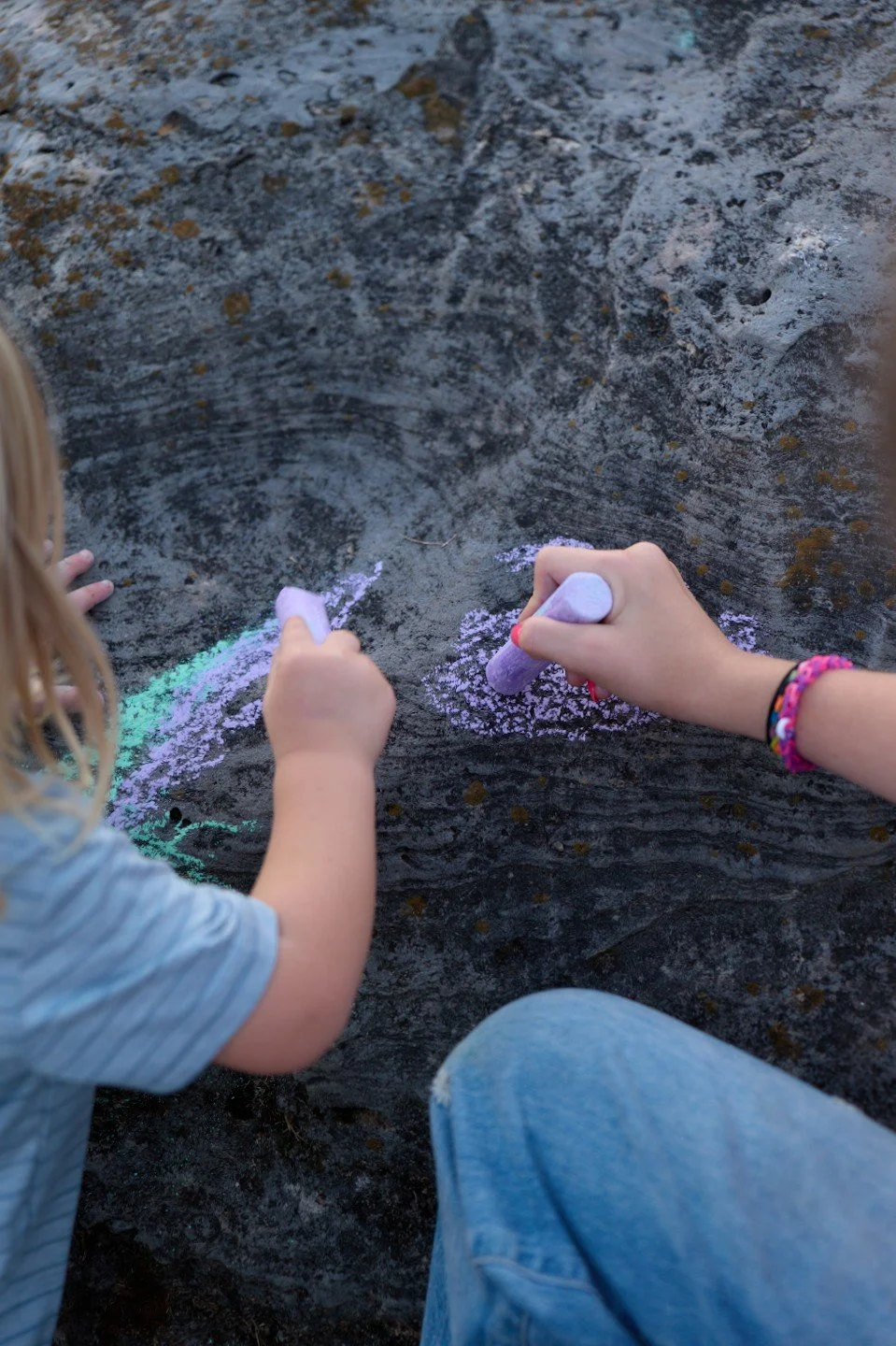 Two children are drawing with pastel chalks on a black surface, possibly a sidewalk or pavement, with visible chalk marks and some colorful light reflecting.