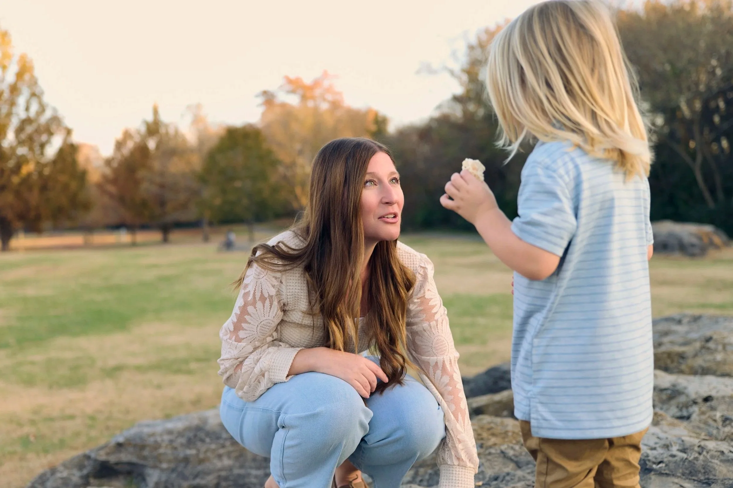 A woman crouching on a rock having a conversation with a young girl in a park during autumn, with trees in the background.