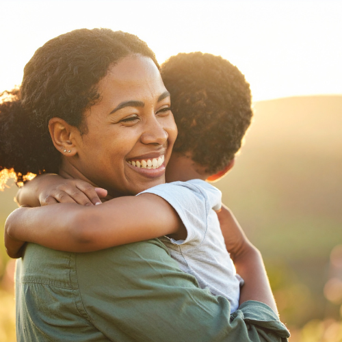 A woman smiling and hugging a child outdoors at sunset.