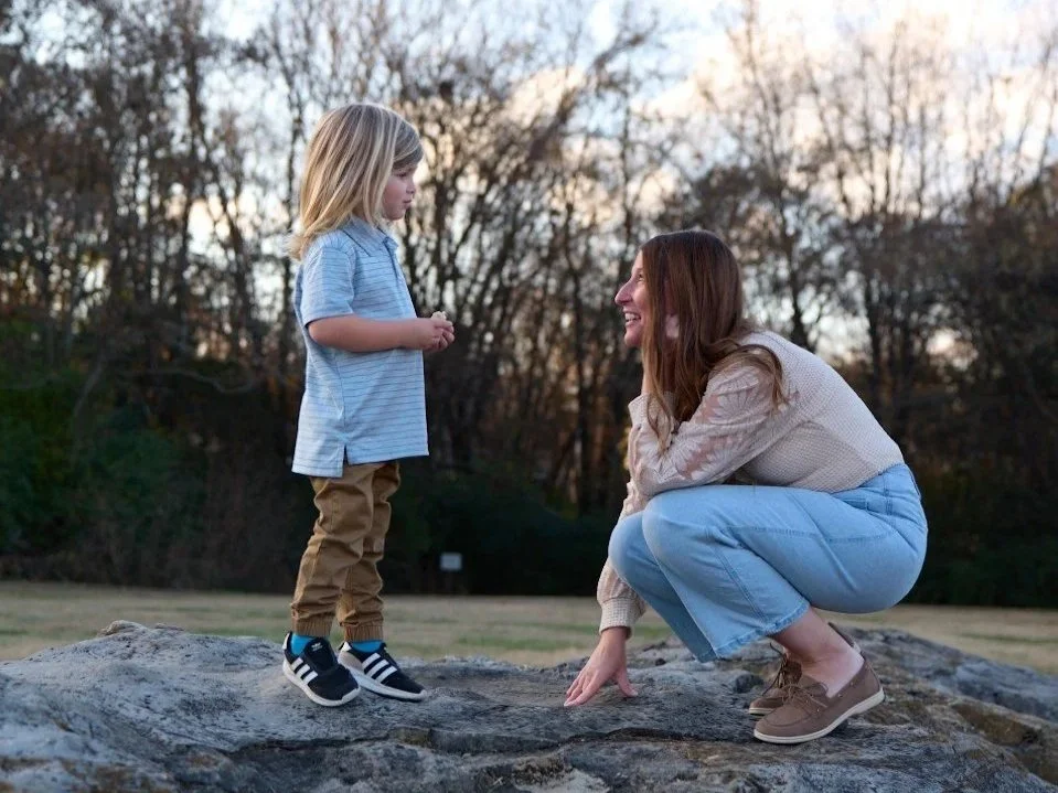 A woman crouching on a rock, smiling at a young girl standing on the rock outdoors during sunset.