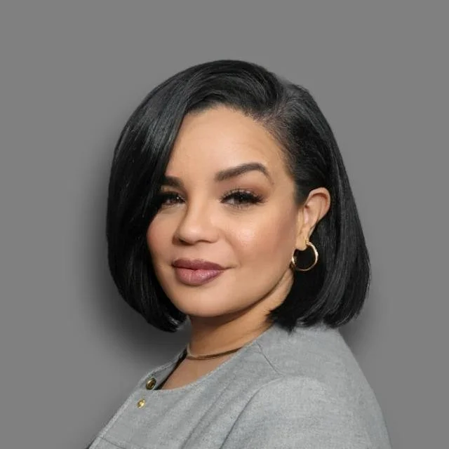 A woman with shoulder-length black hair, wearing hoop earrings and a gray top, posing against a gray background.