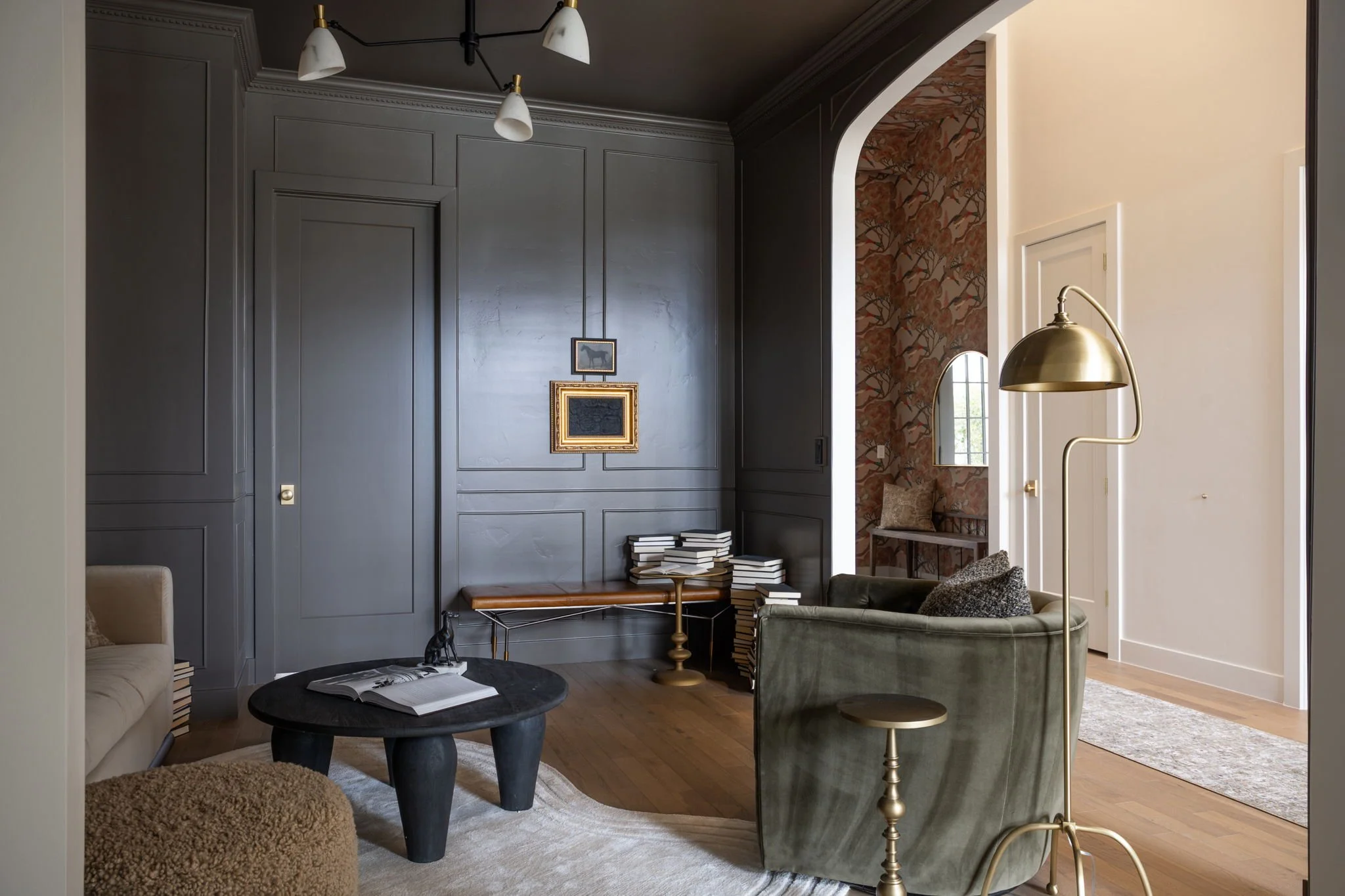 Living room with dark gray paneled walls, a beige sofa, a black coffee table with a book, gold and brass floor lamps, a green velvet armchair with a decorative pillow, a decorative wall with framed art, and a small sitting nook with an arch window an