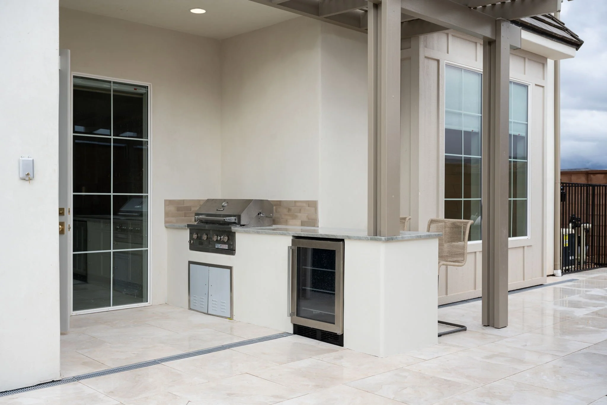Outdoor patio with a stainless steel built-in grill, a mini fridge, and seating area, adjacent to a house with large windows.