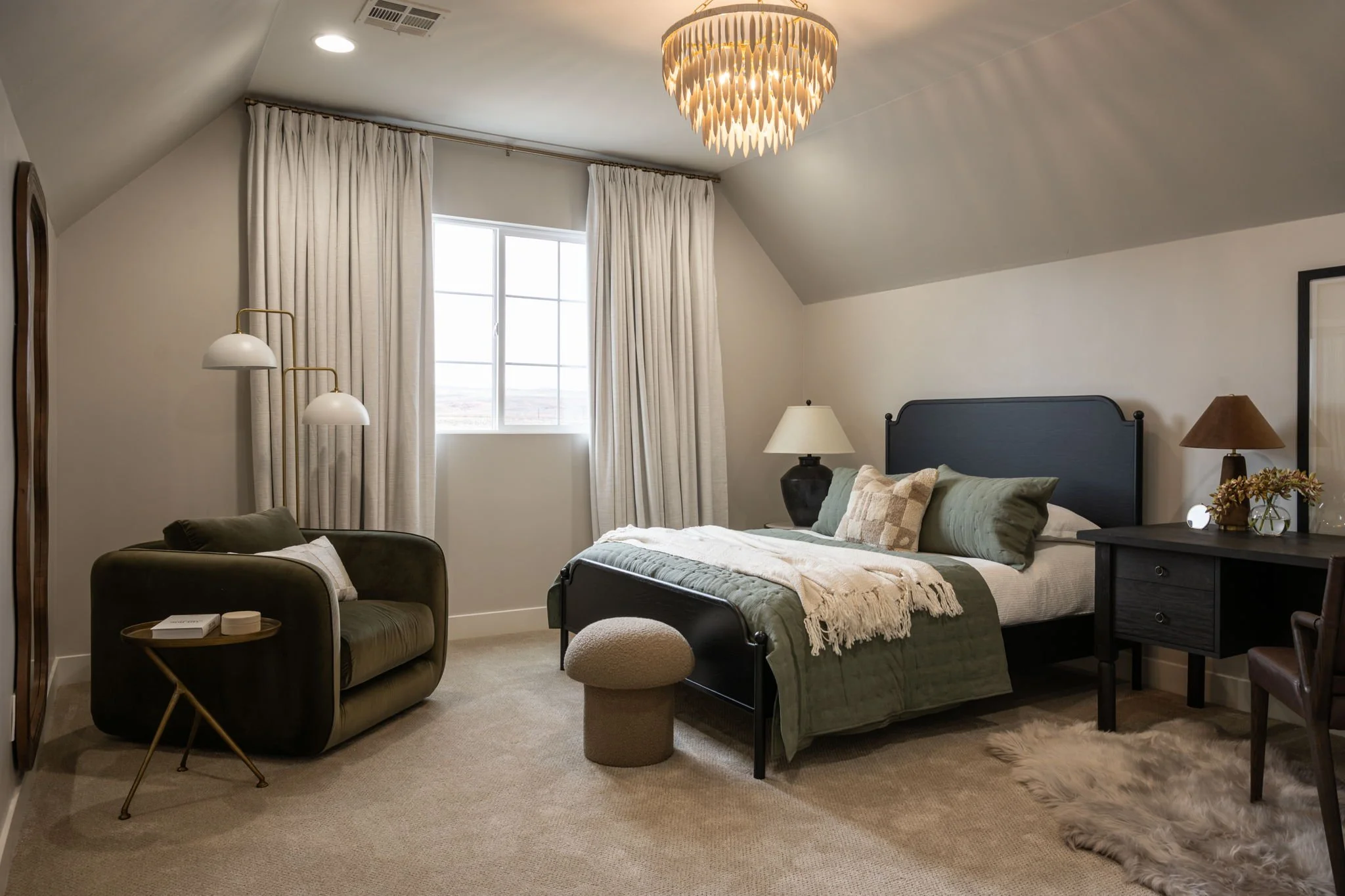 A bedroom with a black metal bed frame, green bedding, and various pillows, next to a black nightstand with a brown lamp. A cream-colored scarf is draped over the bed. To the left, there is a dark green velvet armchair with a small pillow, next to a 