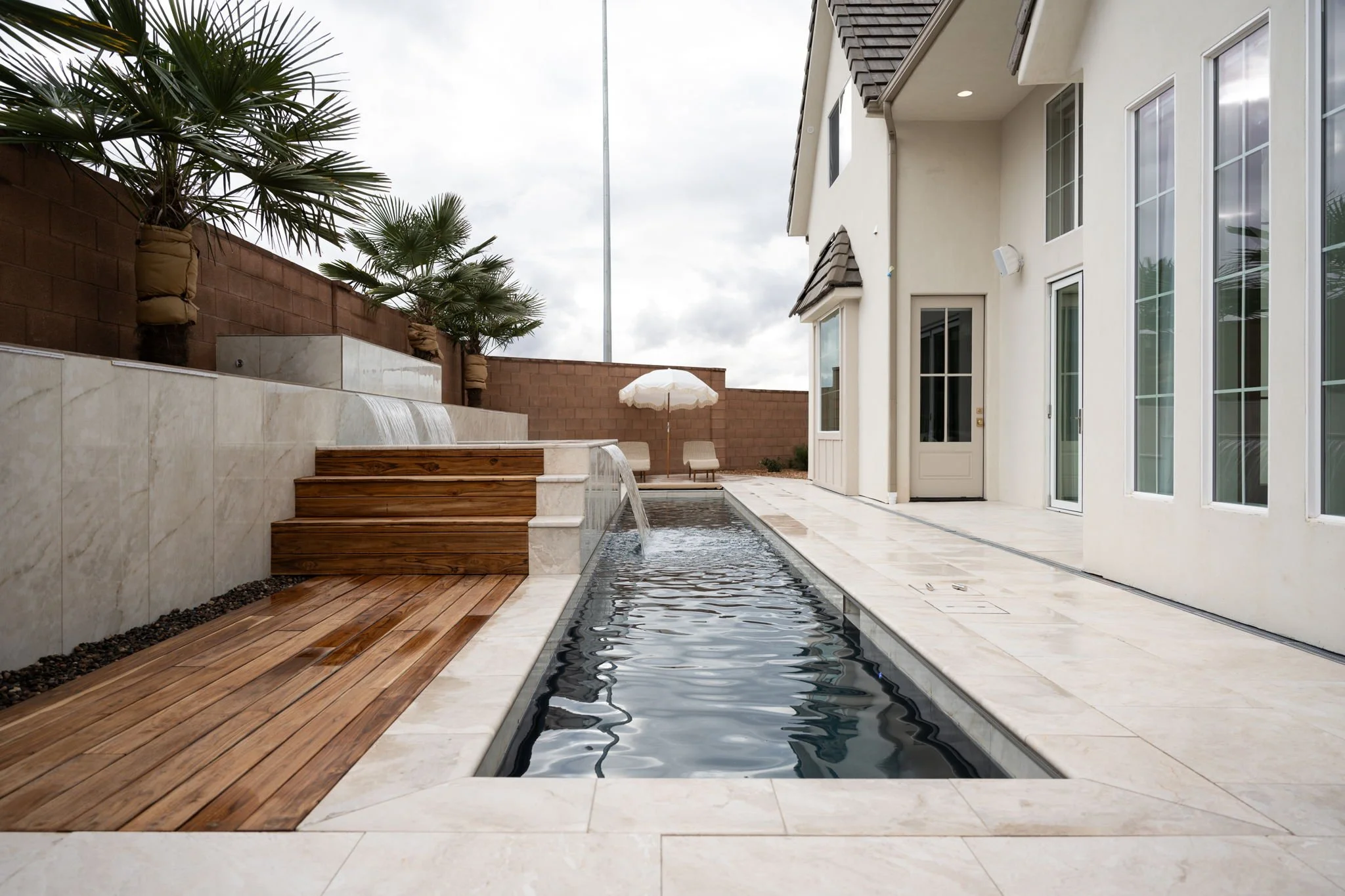 A modern backyard with a narrow rectangular pool, wooden steps, and a water feature with water cascading down a marble wall. There are two lounge chairs with an umbrella at the far end of the pool and several palm trees behind a brick wall. The house