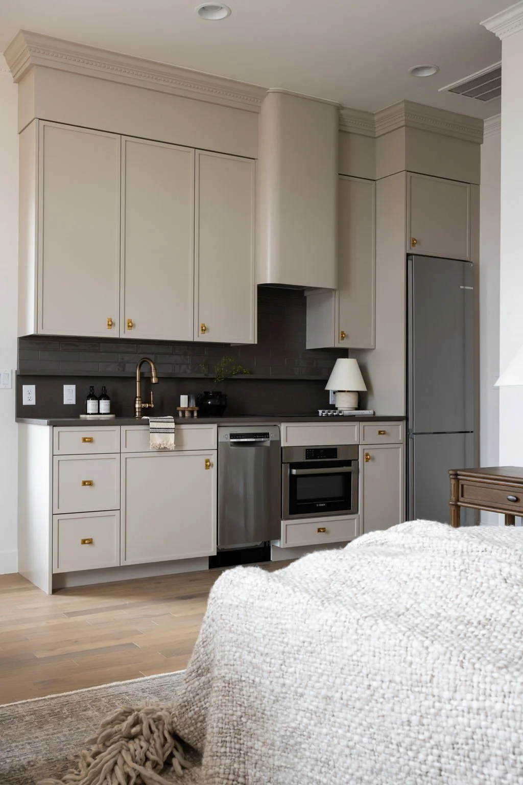 Kitchen with beige cabinets, black backsplash, stainless steel appliances, and wooden flooring, viewed from a living room area with a cream-colored knitted blanket in the foreground.