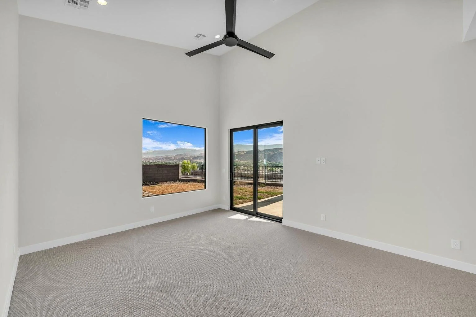 Empty room with beige carpet, white walls, a ceiling fan, a large window, and a sliding glass door leading outside. The view includes mountains and a partly cloudy sky.