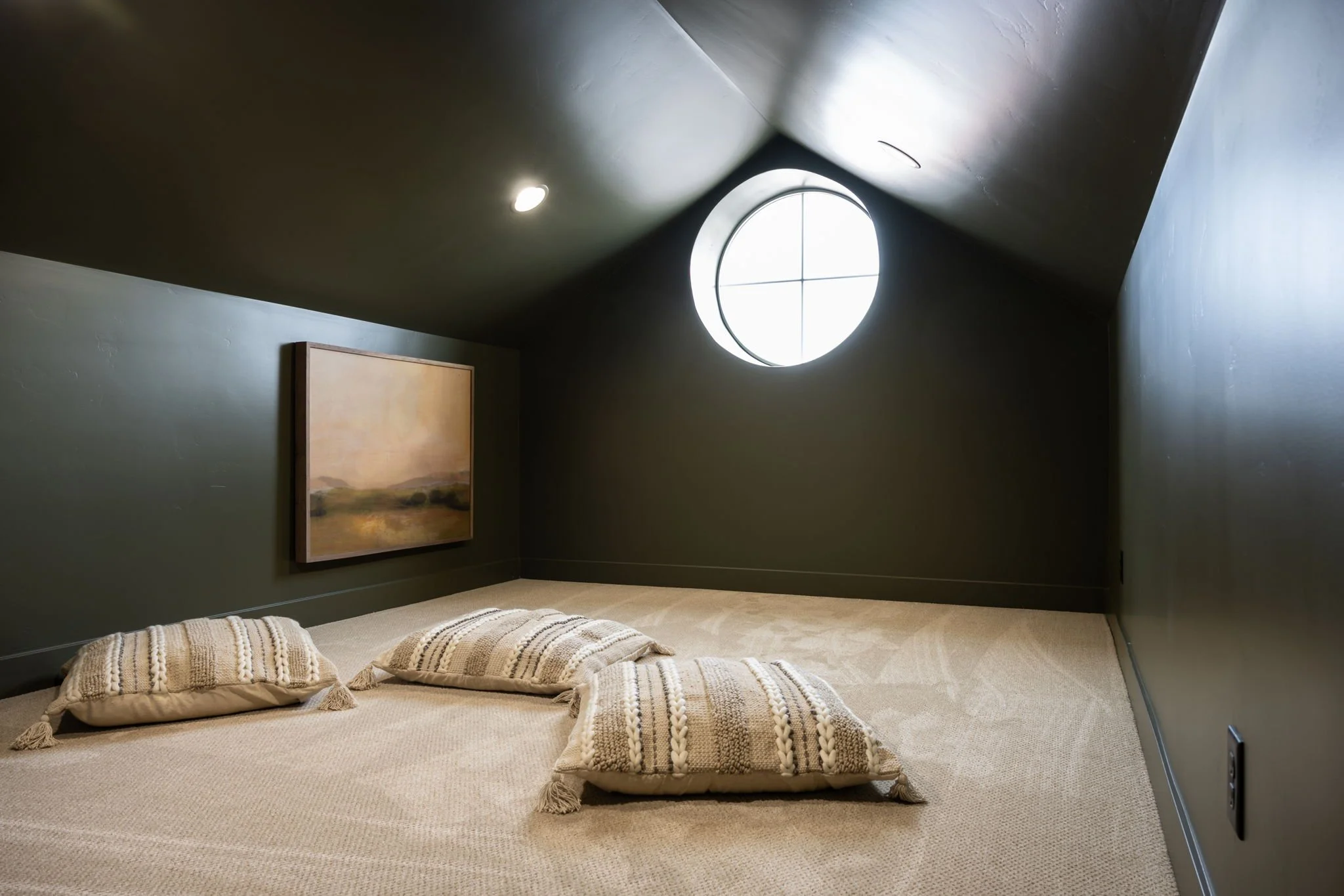 A cozy attic room with dark walls, beige carpet, and a circular window. There are three decorative pillows on the floor and a landscape painting on the wall.
