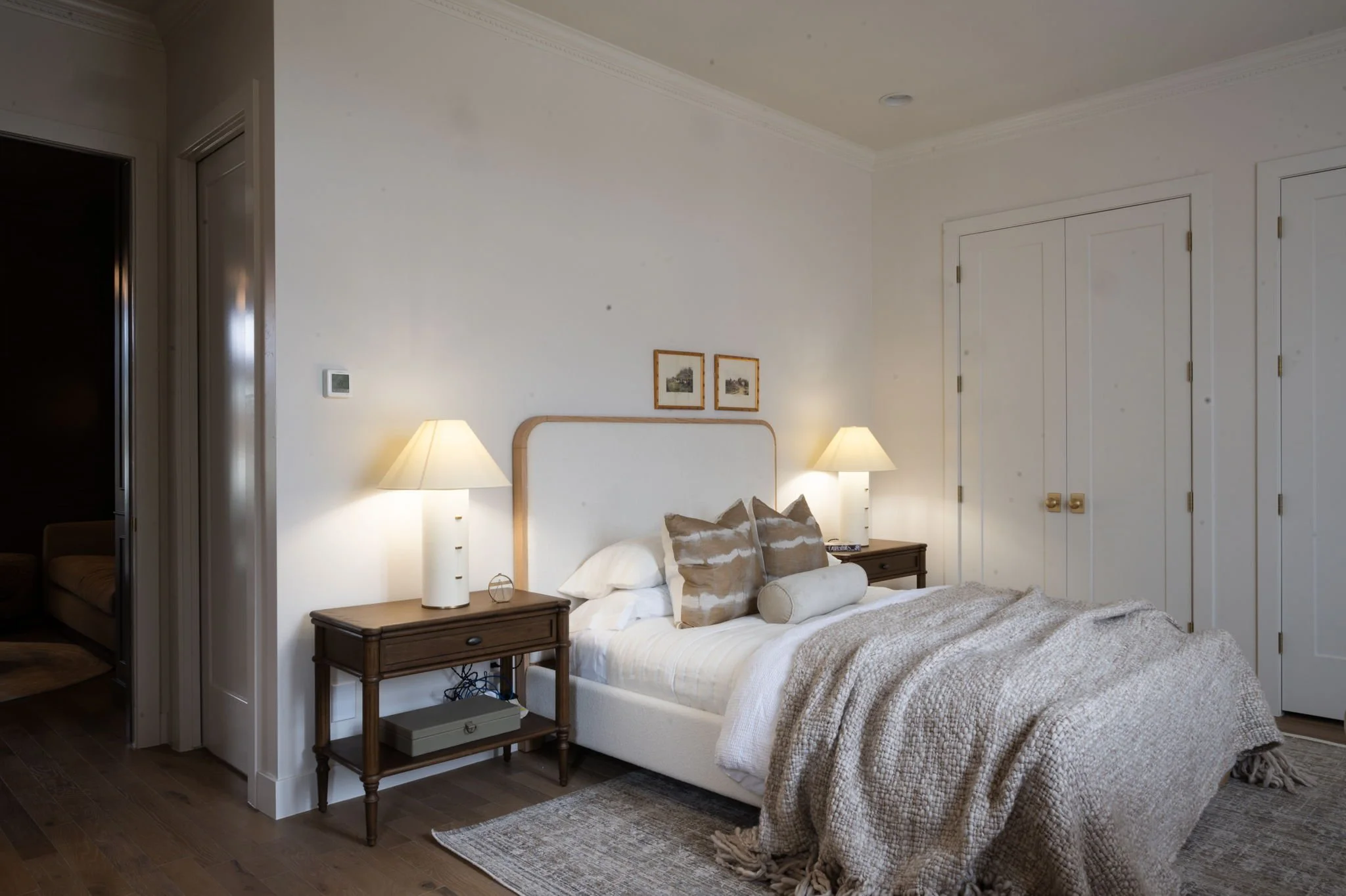 A cozy bedroom with a bed, wooden nightstands, and lamps on each side. The bed has pillows and a textured blanket. There are two small framed pictures above the headboard. The room has white walls, a closet with closed doors, and hardwood flooring.