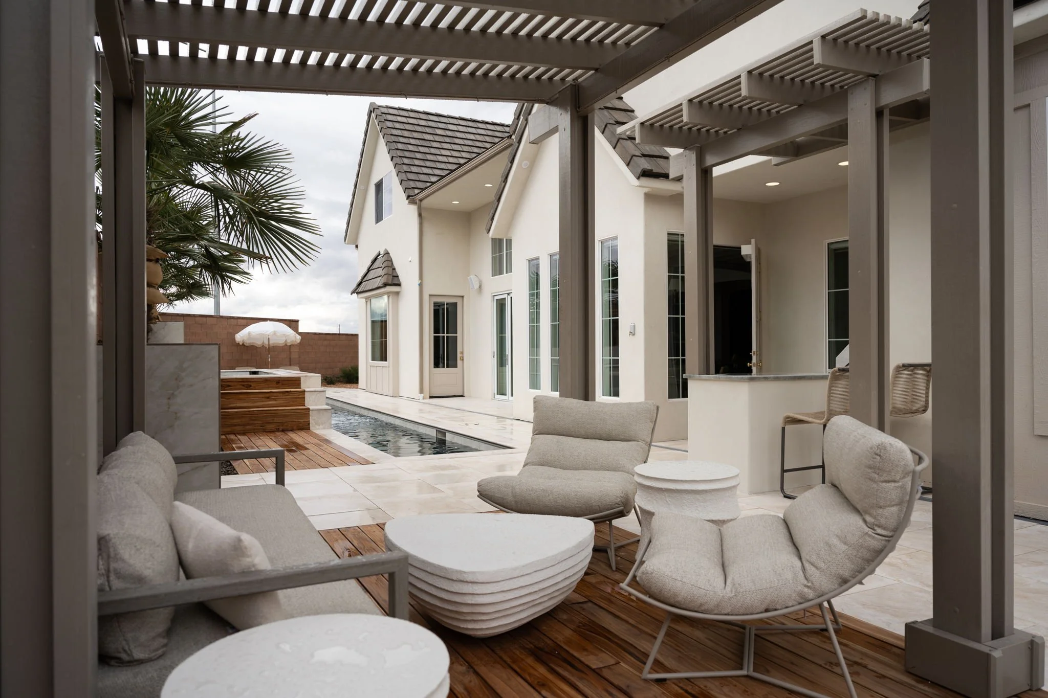 View of a modern backyard patio with outdoor seating, a pool, and a white house with large windows and a tiled roof.