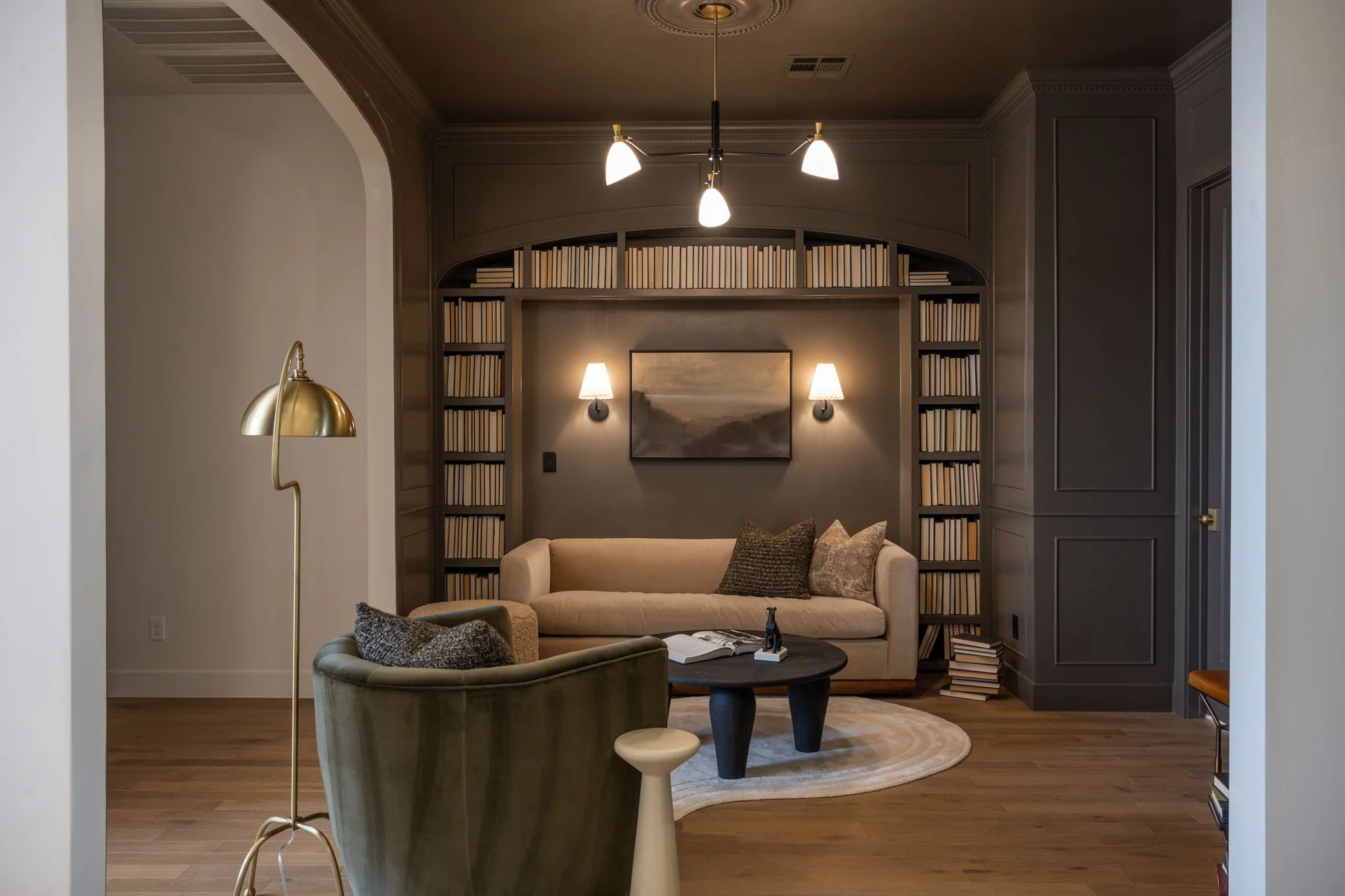 Living room with beige sofa, green armchair, black coffee table, gold floor lamp, and dark wooden built-in shelves filled with books, with a dark wall and framed painting.