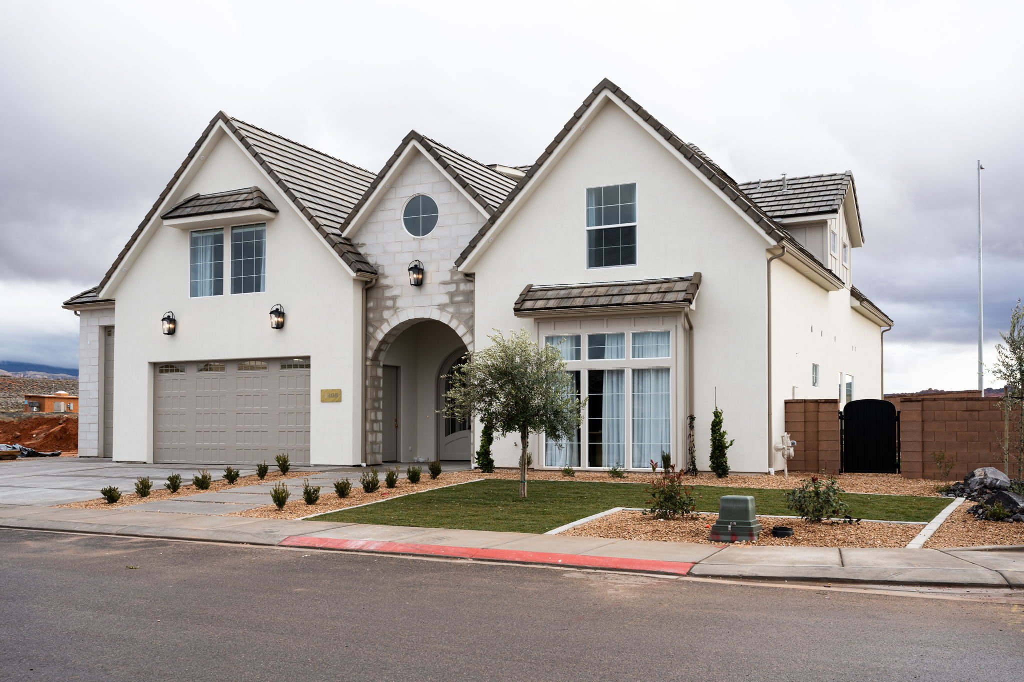 A modern two-story house with white stucco exterior, dark gray roof, and a two-car garage. Features include a front yard with a small tree, shrubs, and a lawn, with a sidewalk and curb in front.