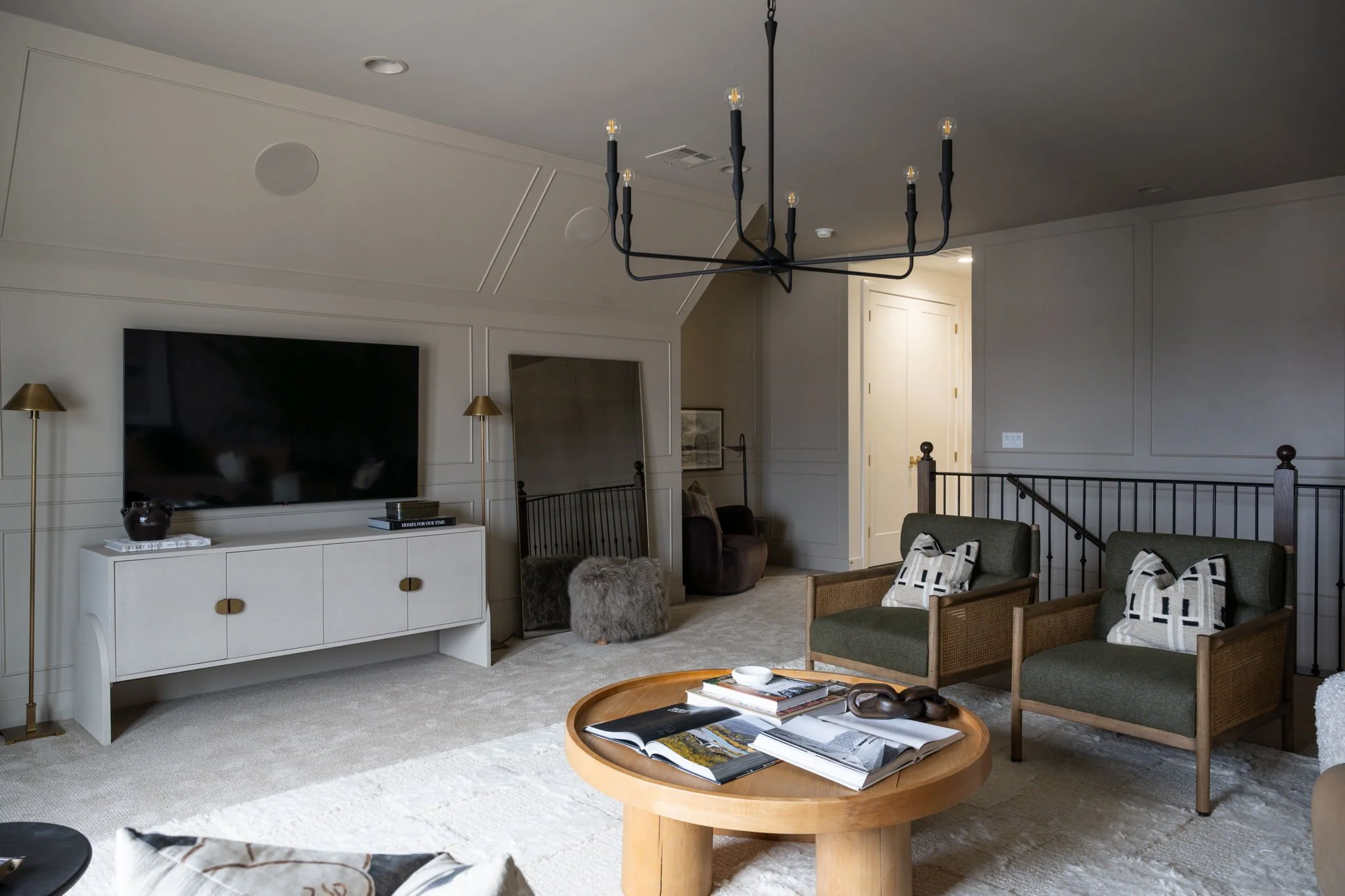 Living room with a wall-mounted flat-screen TV, white TV stand, gold floor and wall lamps, a black chandelier, two green armchairs with patterned pillows, a round wooden coffee table with magazines and a decorative object, mirror, and a small seating