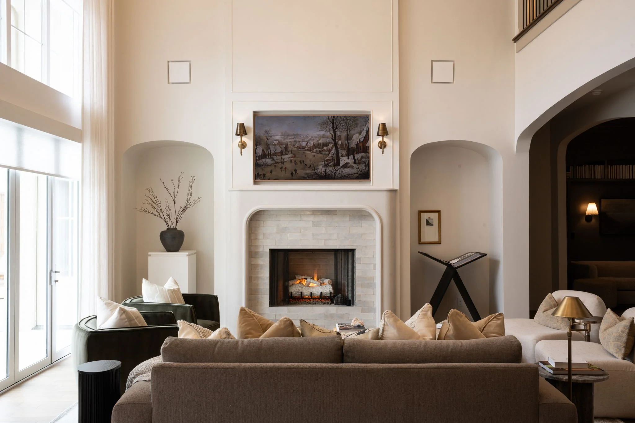 Living room with beige and dark-colored furniture, a fireplace with a white brick surround, wall-mounted lamps, a painting above the fireplace, and decorative items like a vase with branches, a side table with a lamp, and a picture on the wall.