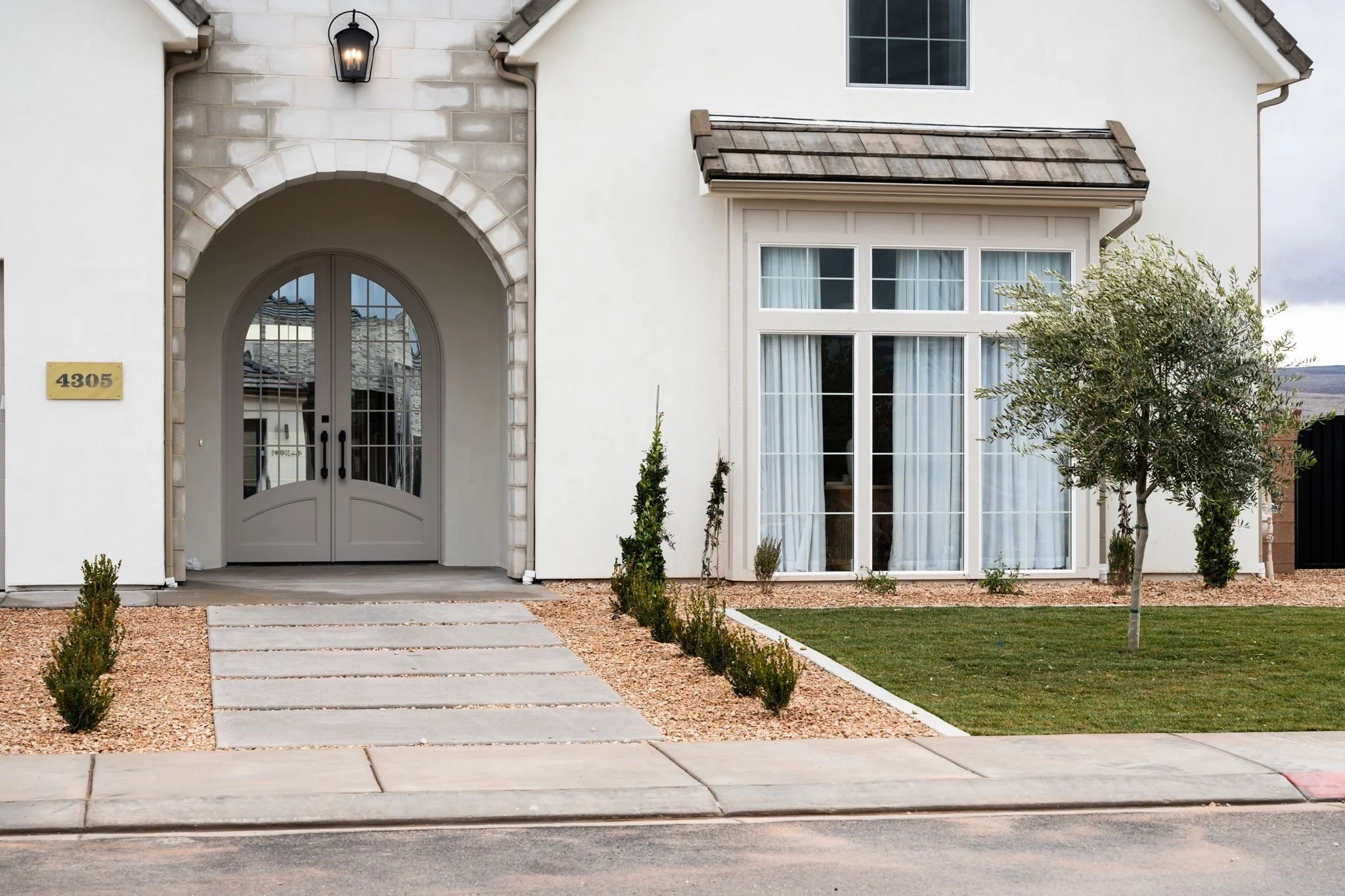 Front view of a modern house with a white exterior, arched front door, and large window with white curtains, small trees and shrubs in the yard, and a sidewalk in the foreground.