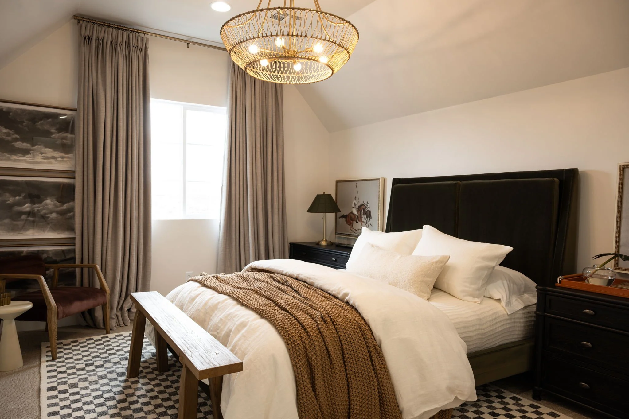 A cozy bedroom with a large bed covered in white linens and a brown knitted throw, black headboard, black nightstands with a lamp and artwork, a bench at the foot of the bed, a window with gray curtains, a chandelier overhead, and a patterned rug on 