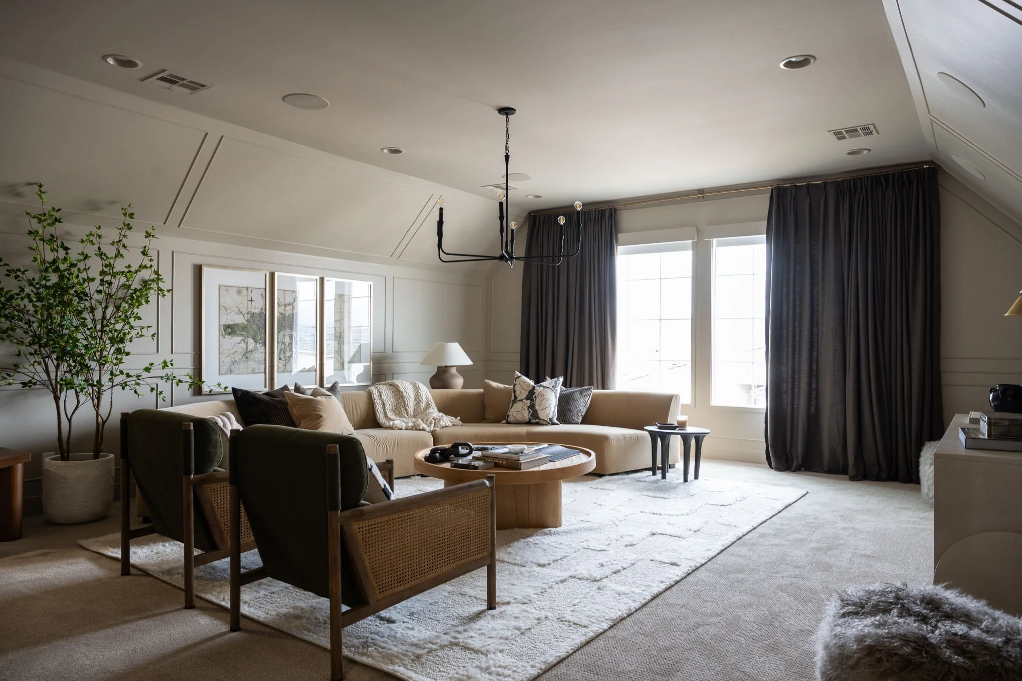 Living room with beige sectional sofa, two dark armchairs, coffee table, large window with dark curtains, white rug, lamp, artwork, and green potted plant.