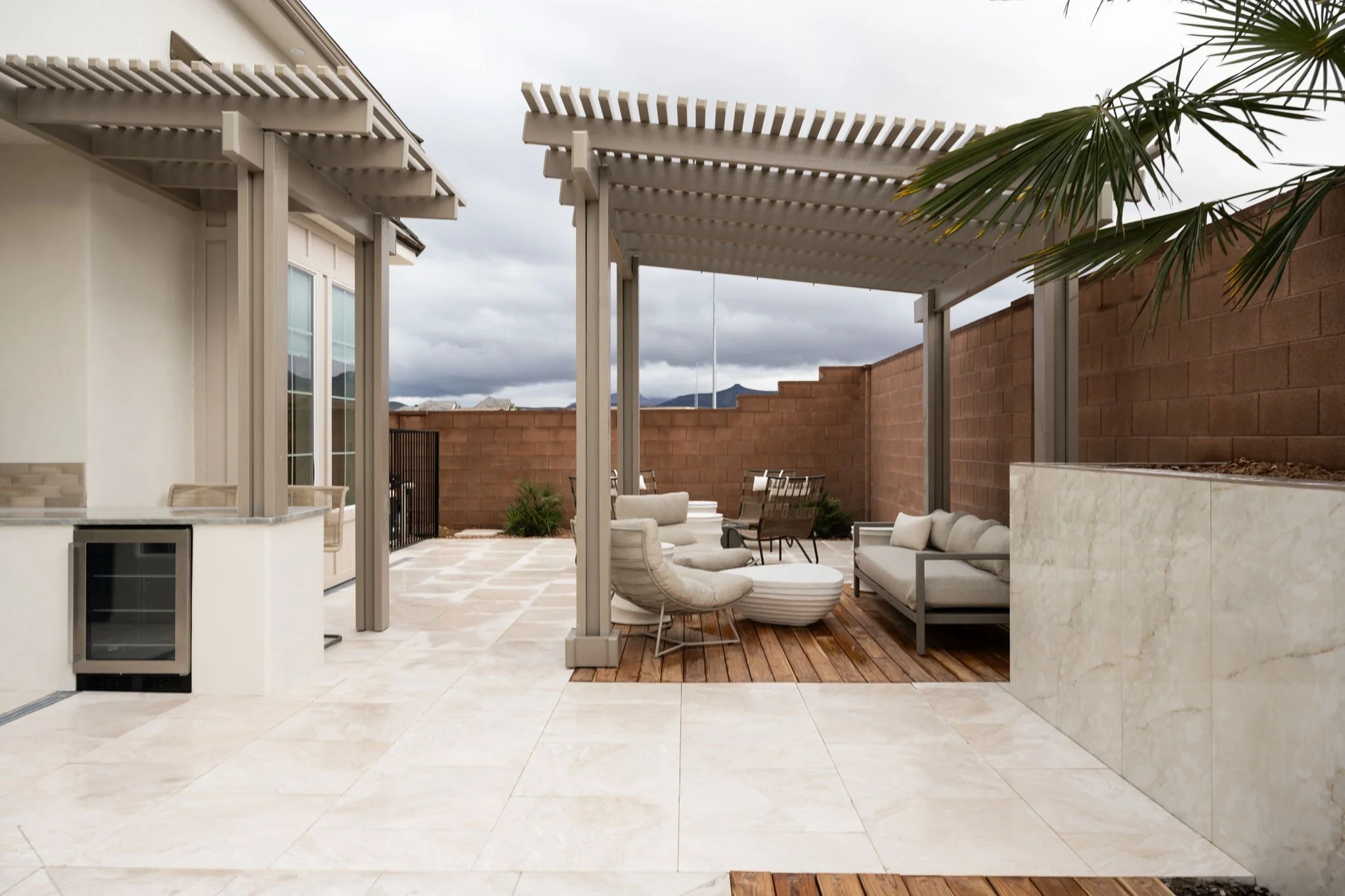 Outdoor patio with seating area under a pergola, beige and white furniture, a wooden deck, and a brick wall in the background with mountains and cloudy sky.
