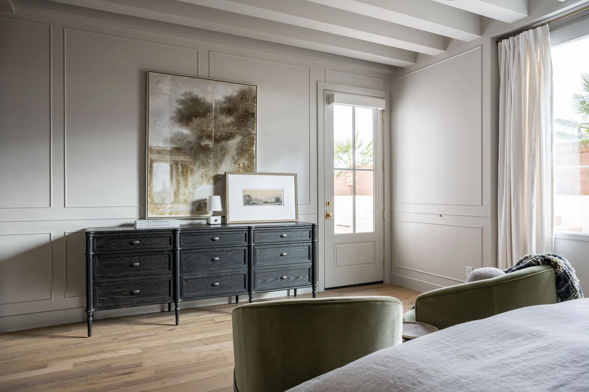 Modern bedroom with light walls, a black wooden dresser, framed artwork, a bedside lamp, and a bed with green chairs nearby. Large windows with white curtains let in natural light.