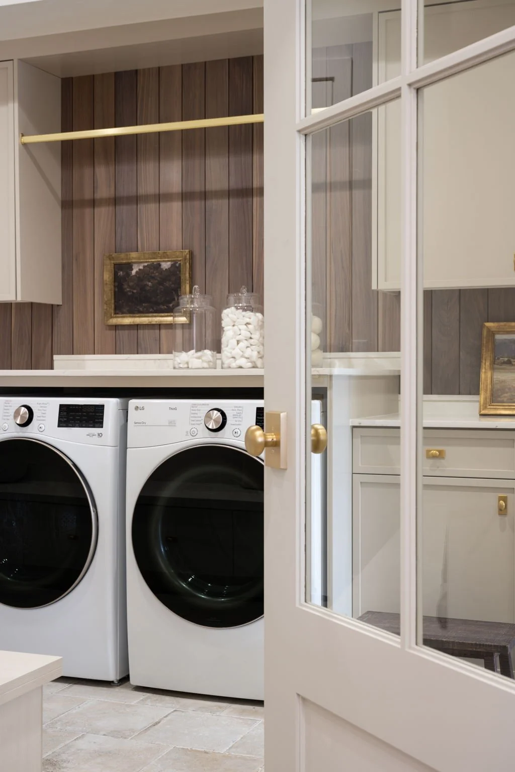 A laundry room with a front-loading washer and dryer, a wooden wall, jars of cotton balls, and a small framed painting.