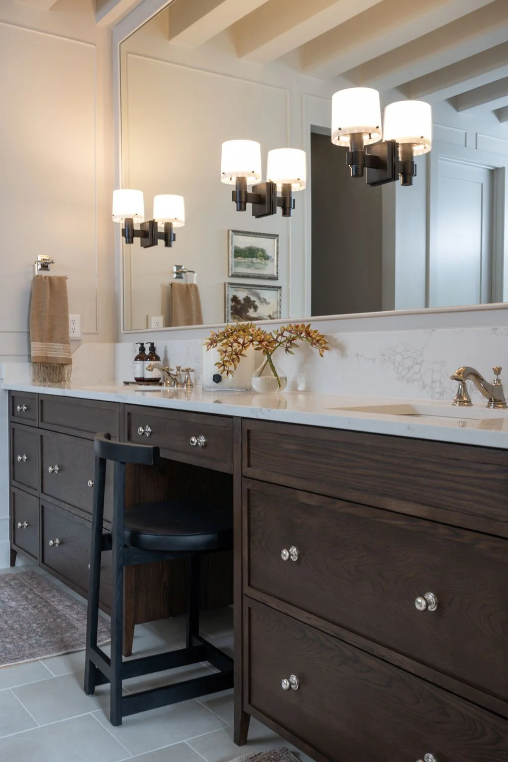 Bathroom with dark wood vanity, white marble countertop, large mirror, black and white light fixtures, beige towels, decorative paintings, and a vase with flowers.