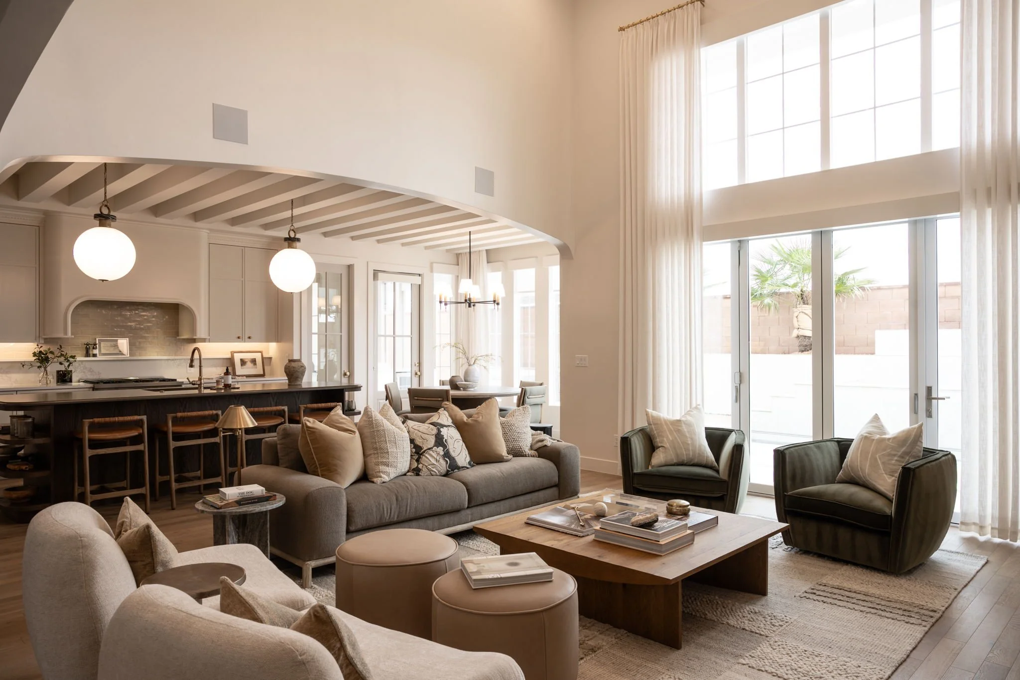 Bright living room with large windows, beige sofa, green armchairs, wooden coffee table, and kitchen in the background with bar stools, pendant lights, and a dining area.