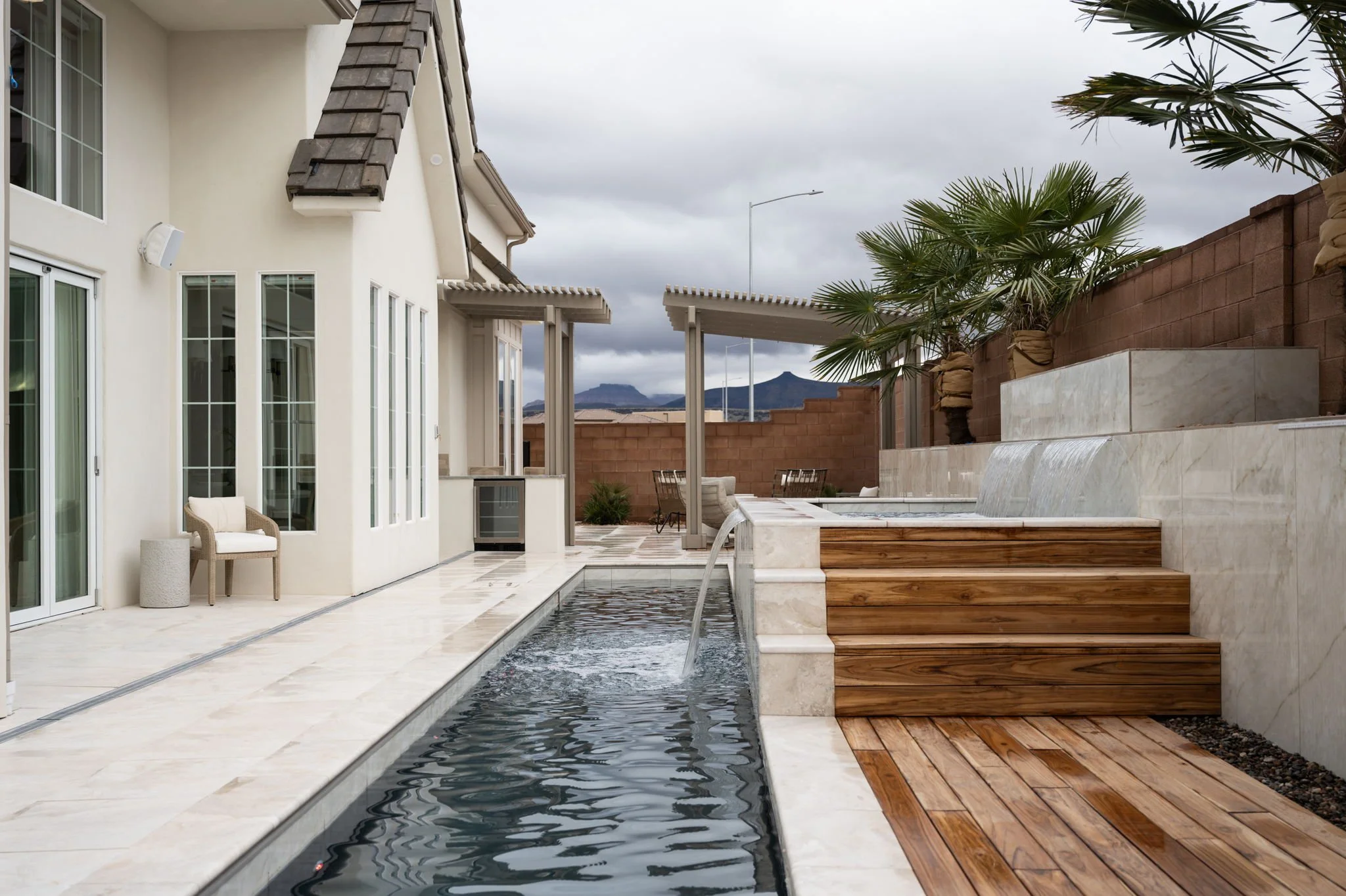 Outdoor backyard with a swimming pool, wooden steps, palm trees, patio furniture, and a house with large windows and sliding doors.