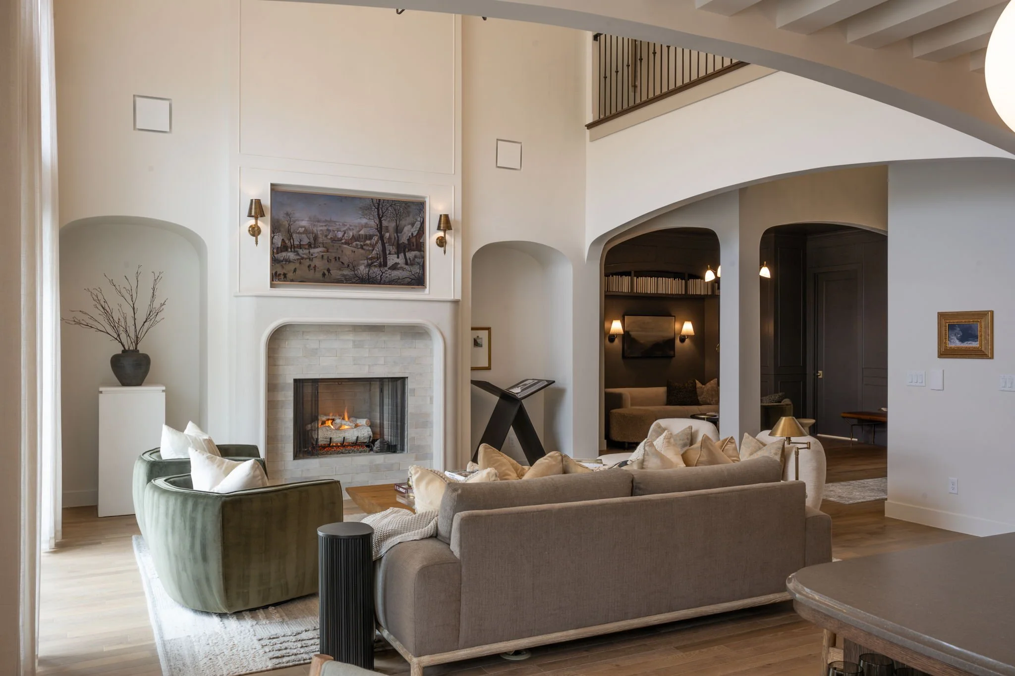 Living room with a beige sofa, a green armchair, a fireplace, and a high ceiling with curved architecture. Artwork hangs above the fireplace, and various lamps provide lighting. There is a vase with branches on a stand to the left and a small framed 