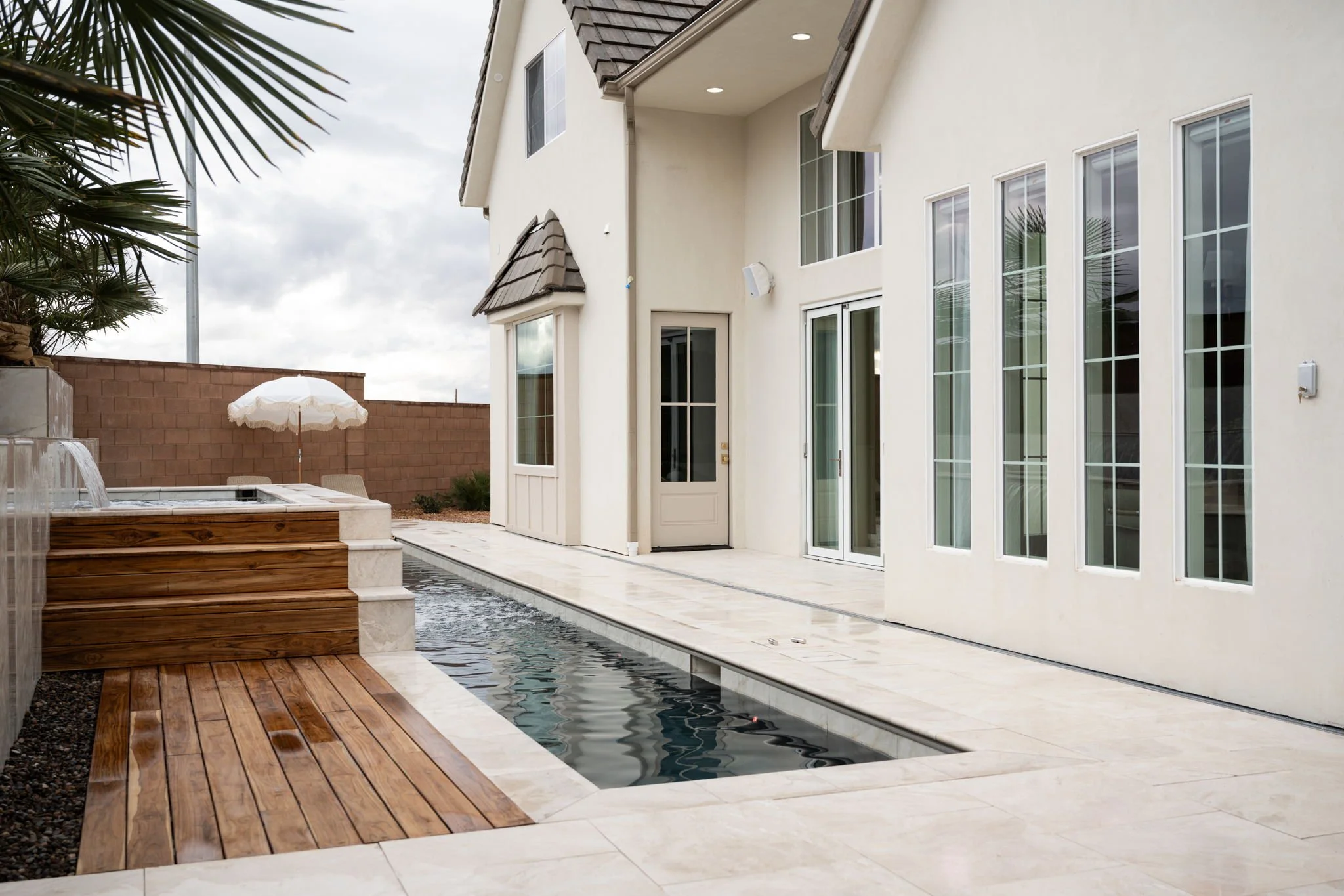 Backyard with a small swimming pool, wooden deck, patio area, and white house with multiple windows and sliding doors.
