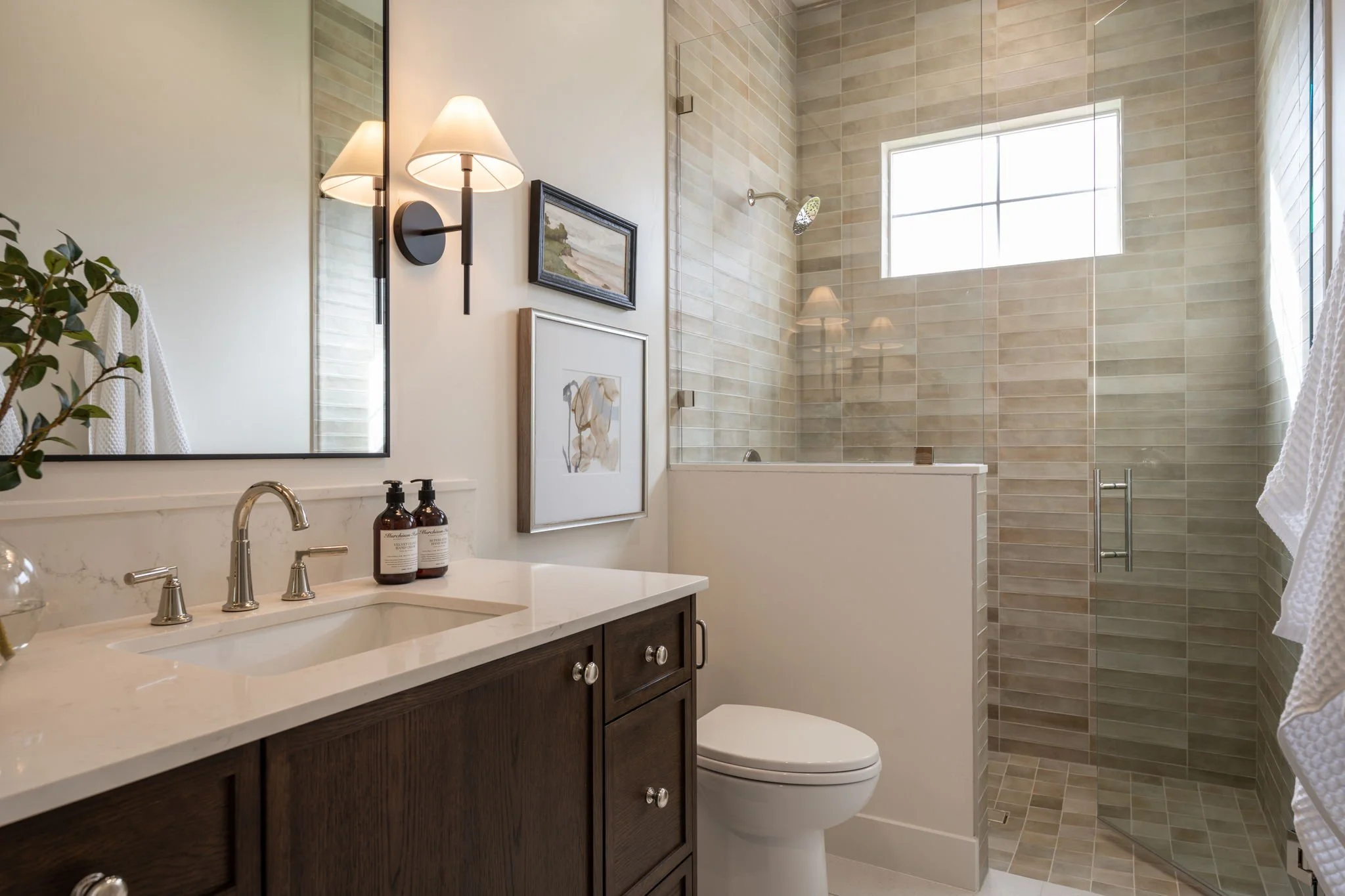 Modern bathroom with a dark wood vanity, white countertop, and a large mirror. Wall-mounted light fixtures, framed artwork, a potted plant, and a soap dispenser are visible. A walk-in shower with beige tiles and a window are in the background.
