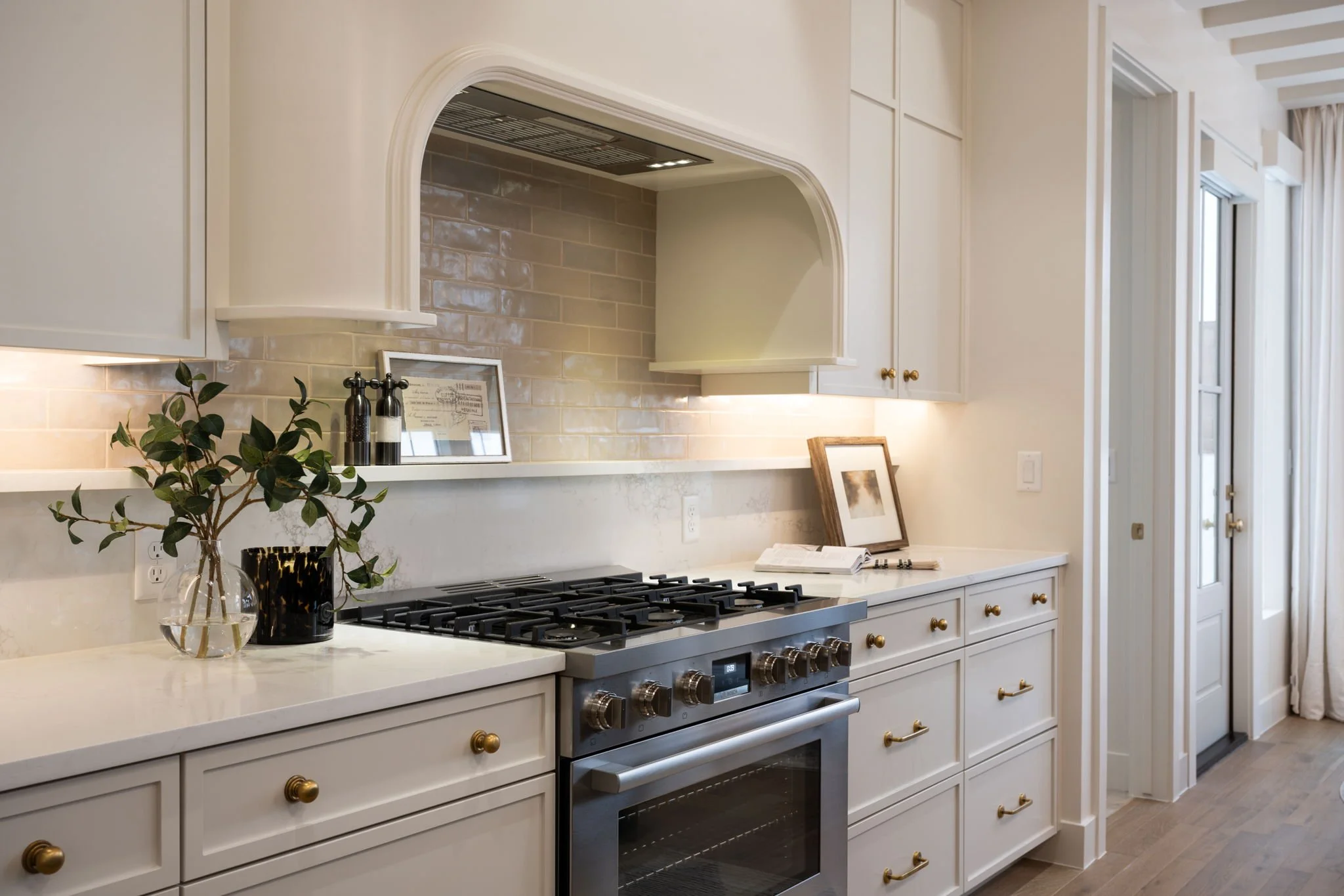 A modern kitchen with white cabinets, a beige backsplash, a stainless steel oven, and a countertop with decorative plants and framed pictures.