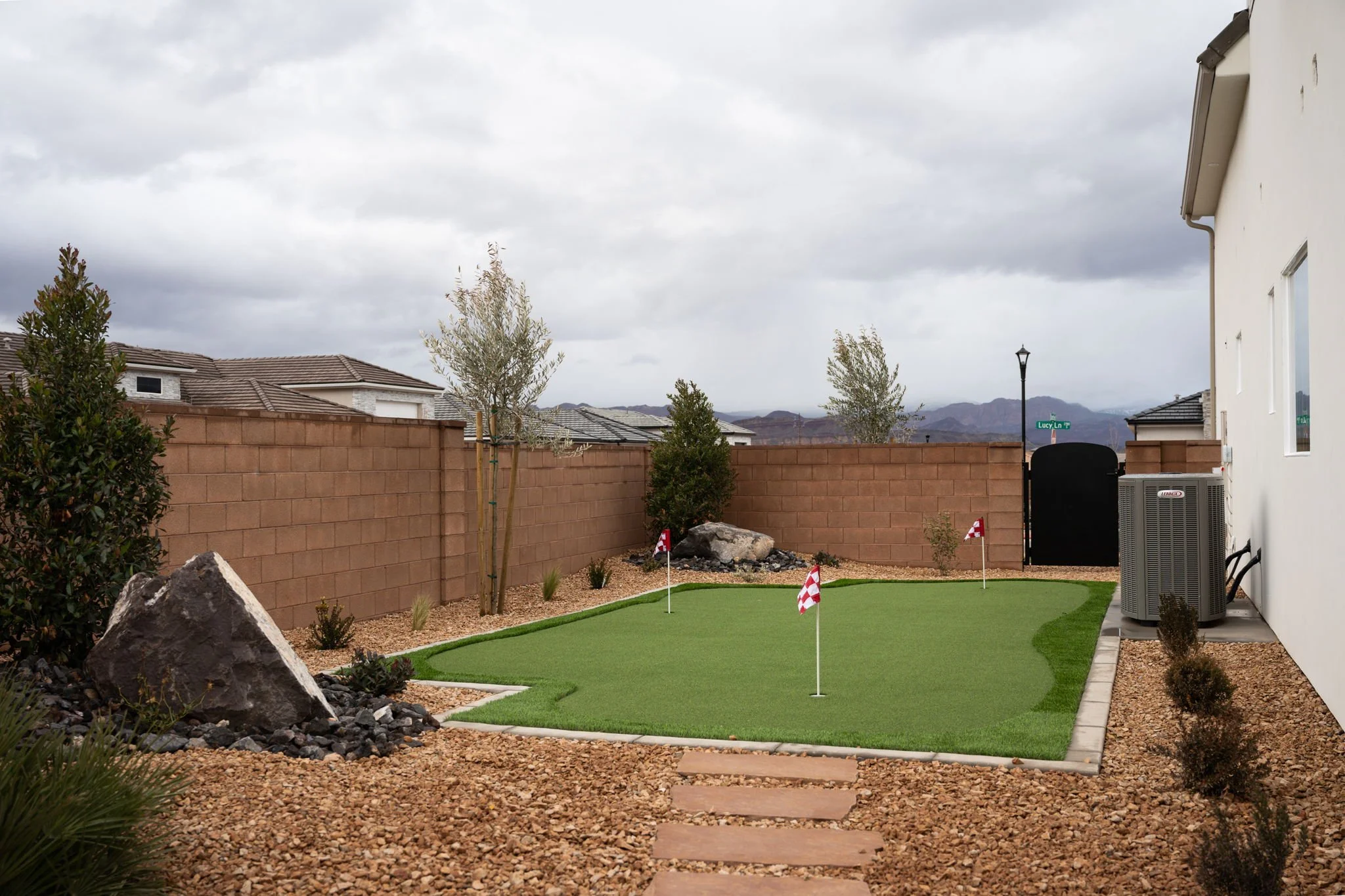 Backyard with a small putting green, flags, small trees, decorative rocks, and a brick wall, under a cloudy sky.
