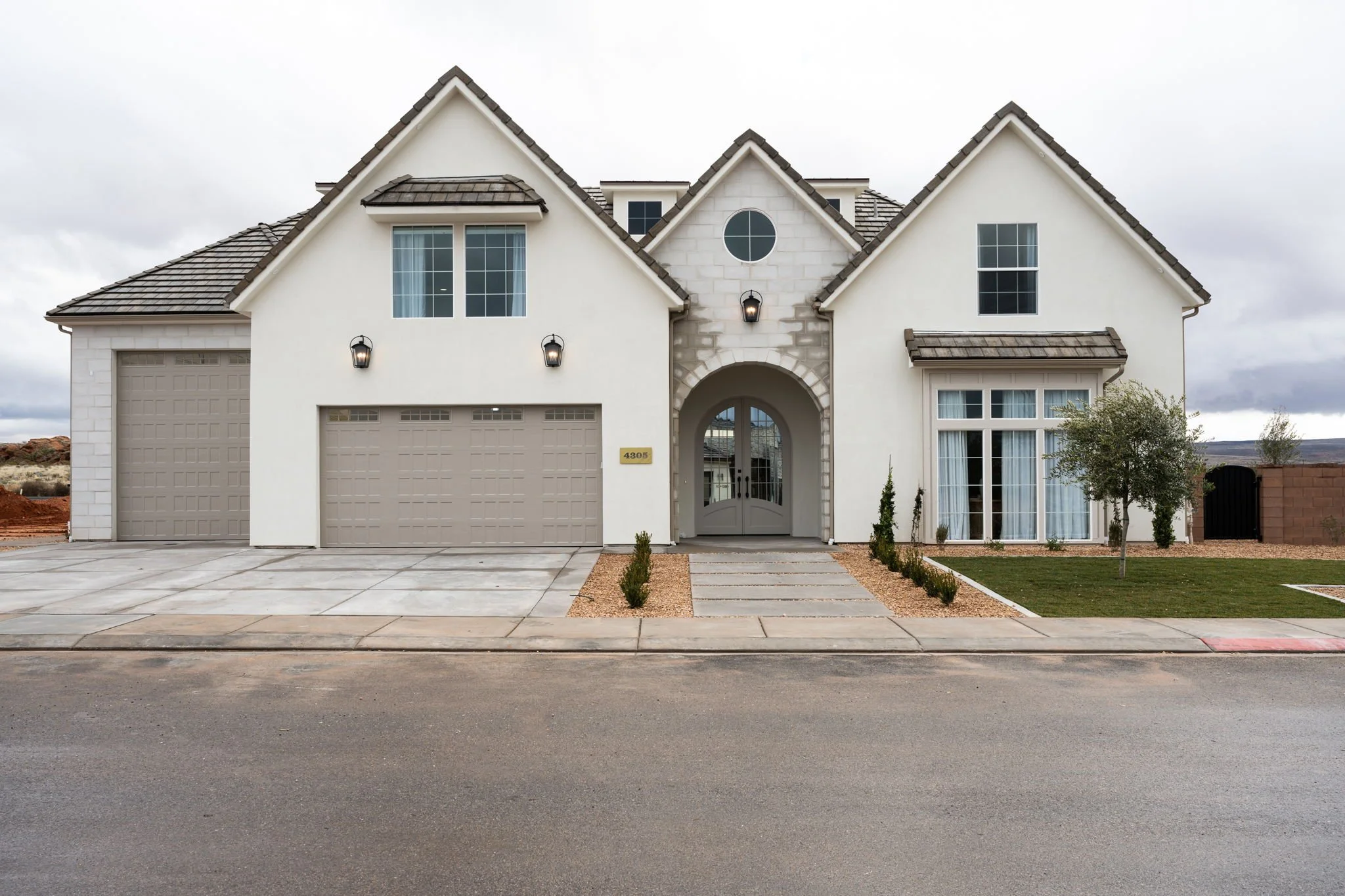Front view of a modern two-story house with a beige driveway, a manicured lawn, small trees, and a gray sidewalk. The house has white walls, gray roof, and features a large arched entryway, two garage doors, and multiple windows.
