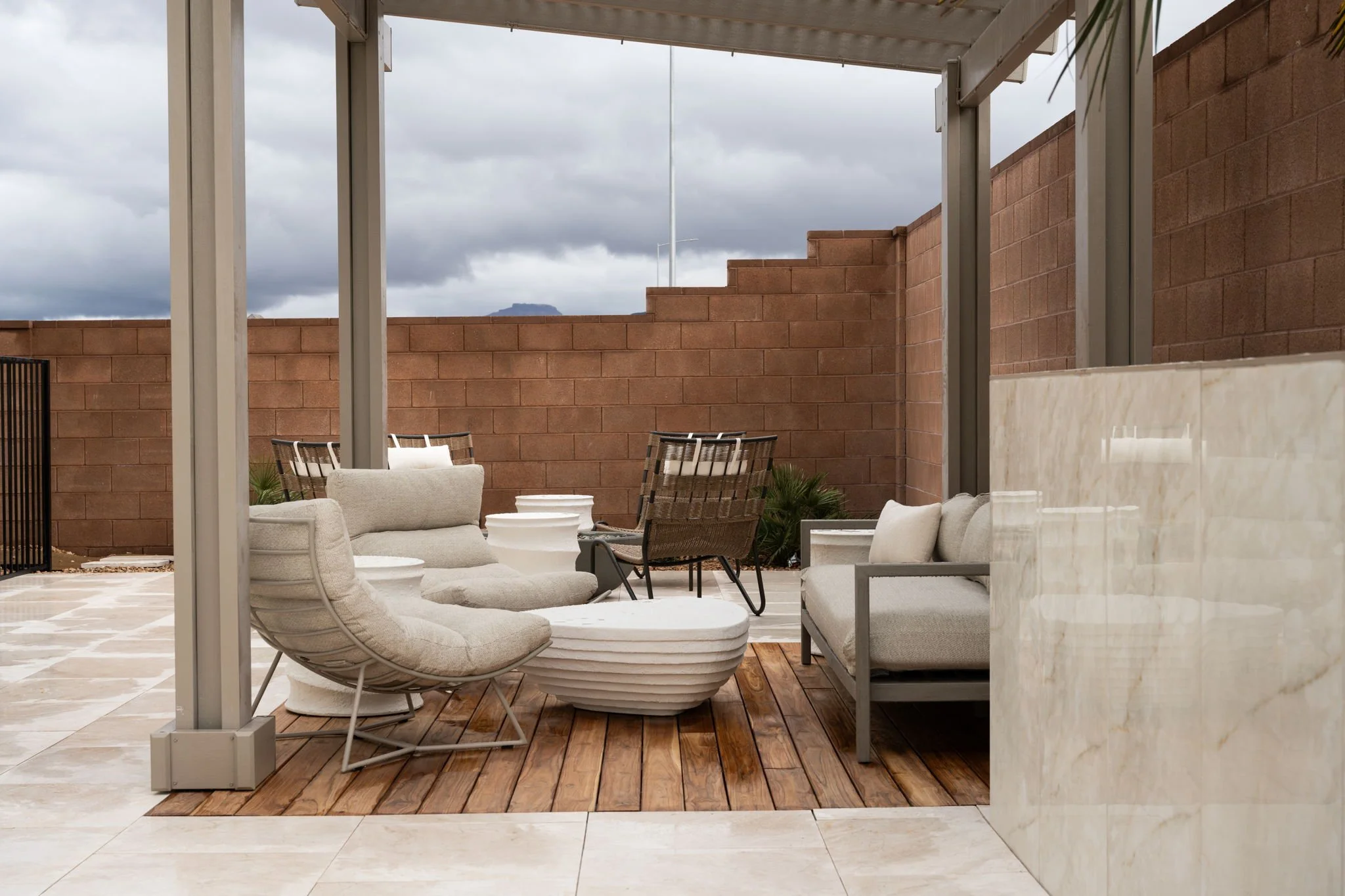 An outdoor patio with modern seating including cushioned chairs and a sofa, a wooden deck area, and a brick wall with a cloudy sky in the background.